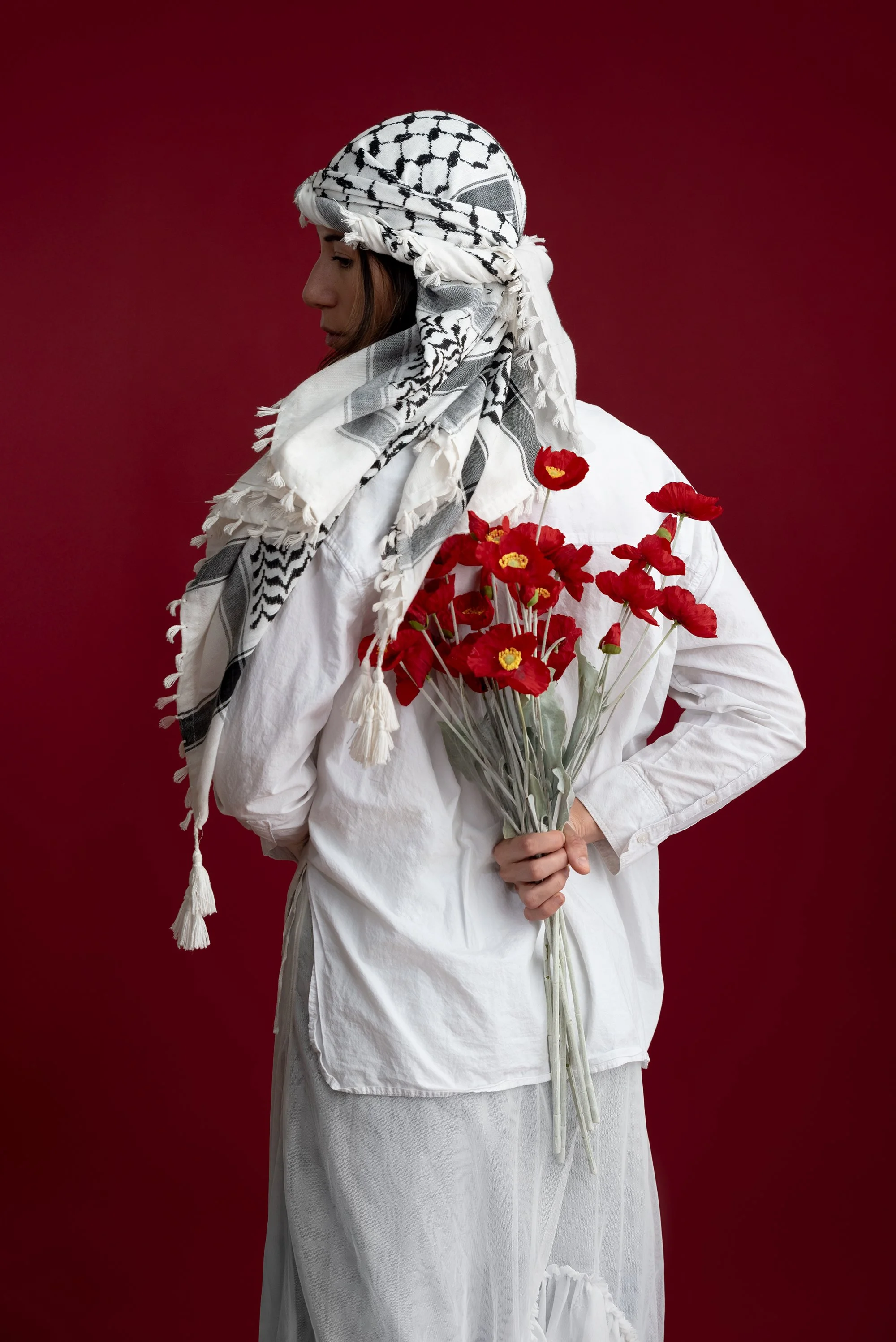 A person wearing a black and white keffiyeh headscarf, dressed in a white shirt and skirt, holding a bouquet of red poppies against a maroon background.