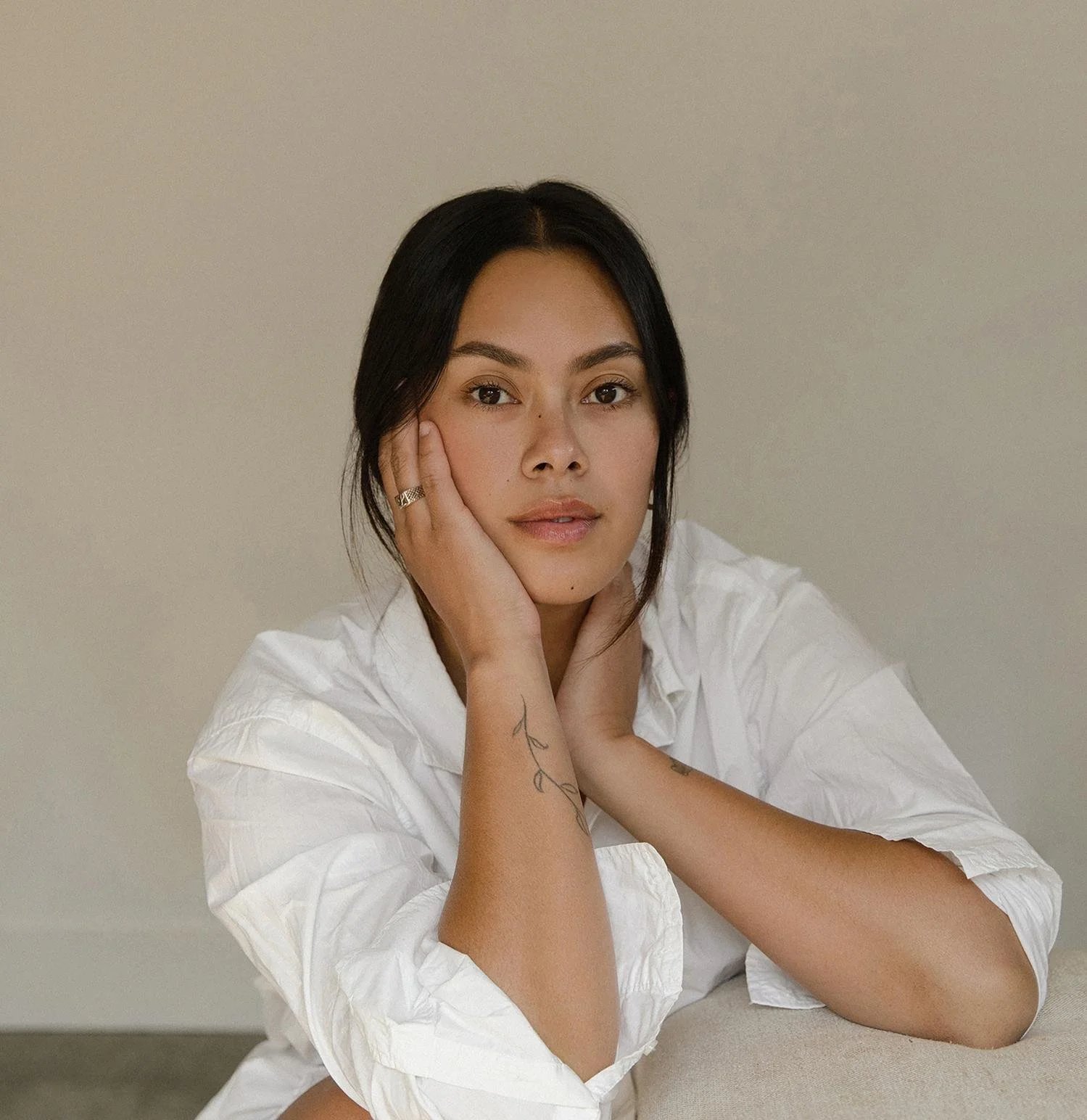 A young woman with black hair, wearing a white shirt, resting her face on one hand while looking at the camera, with a neutral background.