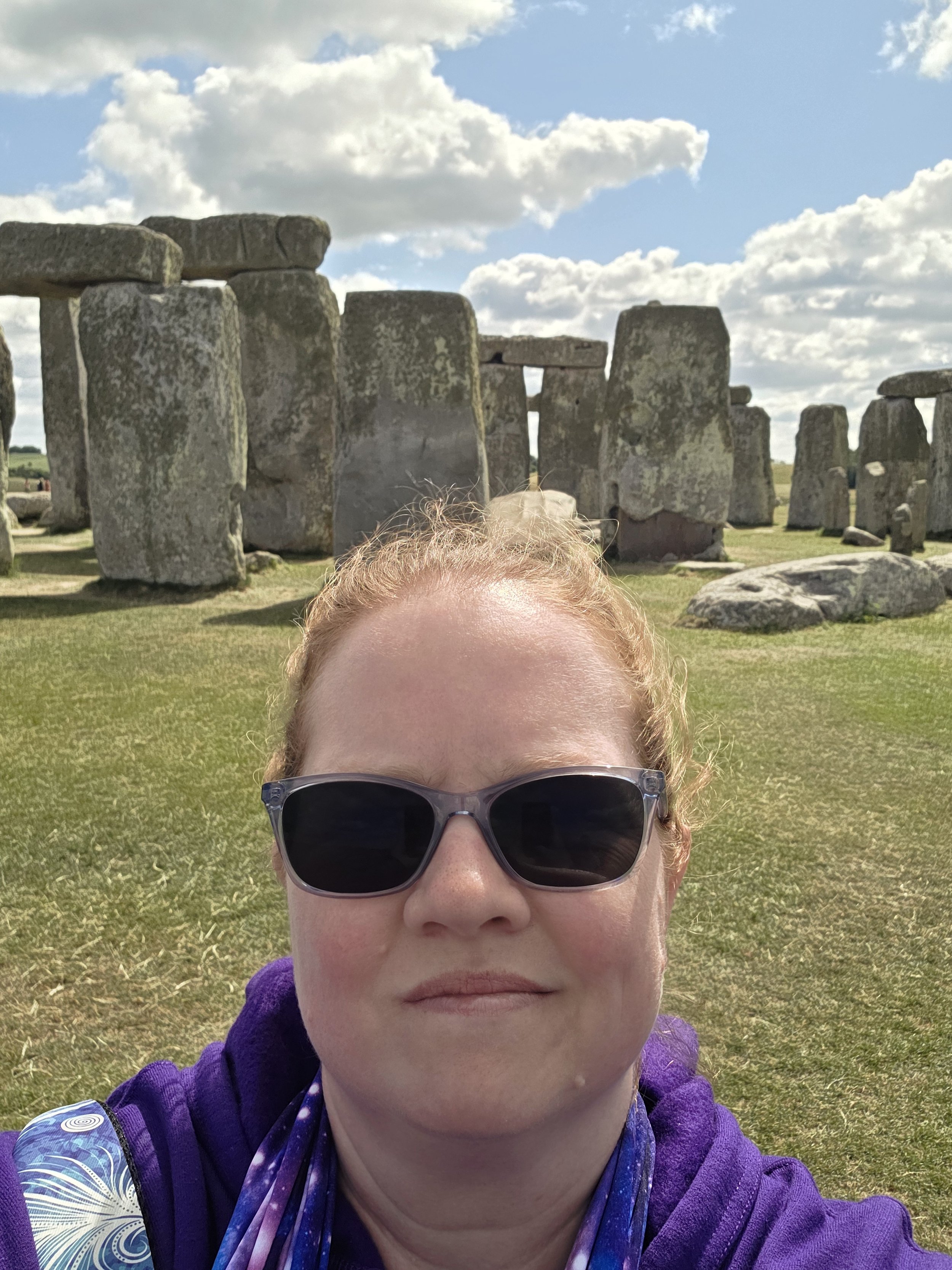 Person wearing sunglasses, purple jacket, and scarf, taking a selfie in front of Stonehenge under a partly cloudy sky.