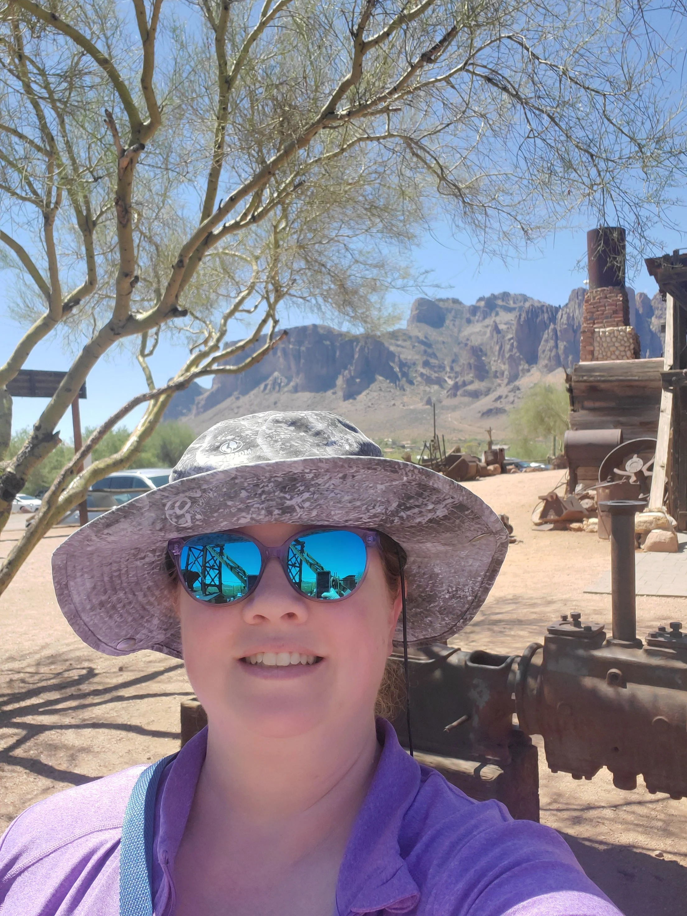 A woman taking a selfie outdoors at a historic desert site with mountains in the background, wearing a sun hat and sunglasses.