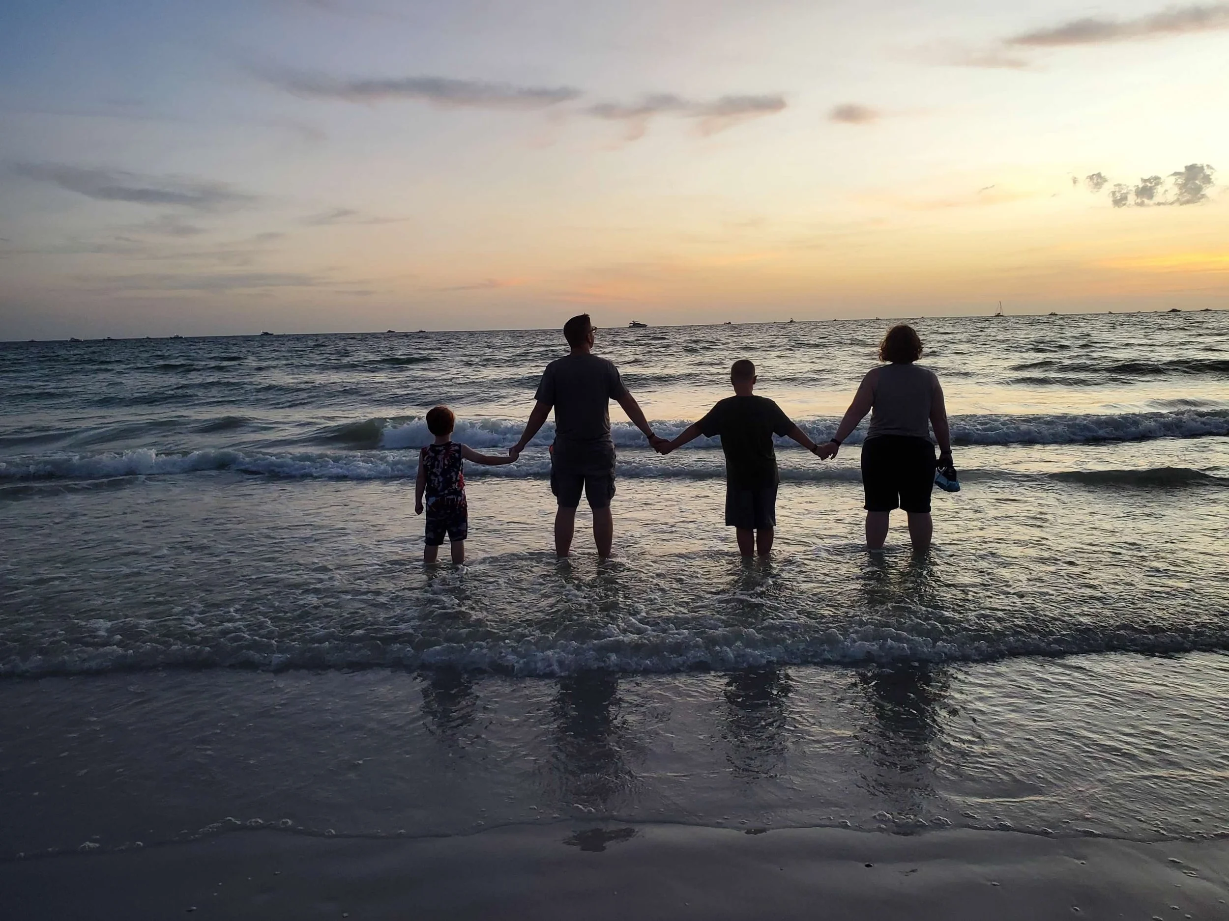 A family of four holding hands in the shallow surf on a beach at sunset, with boats on the horizon.