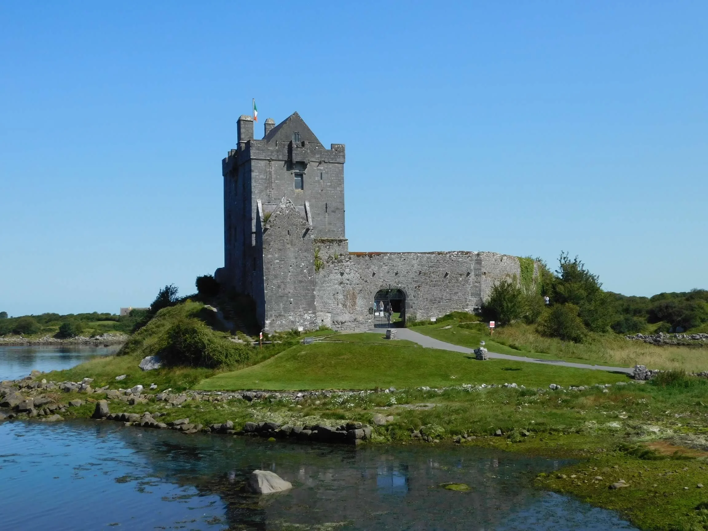 Stone medieval castle with an Irish flag beside a calm body of water under a clear blue sky