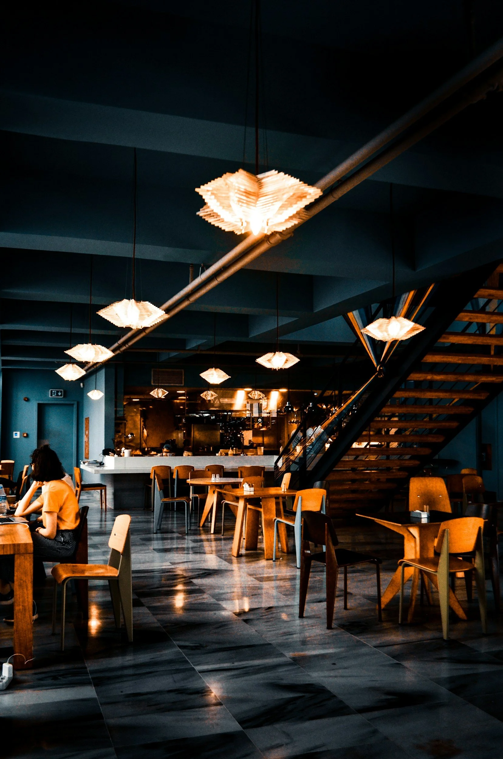 Interior of a modern restaurant or cafe with wooden tables and chairs, some illuminated pendant lights on the ceiling, a staircase on the right side, and a person sitting at a table using a laptop.