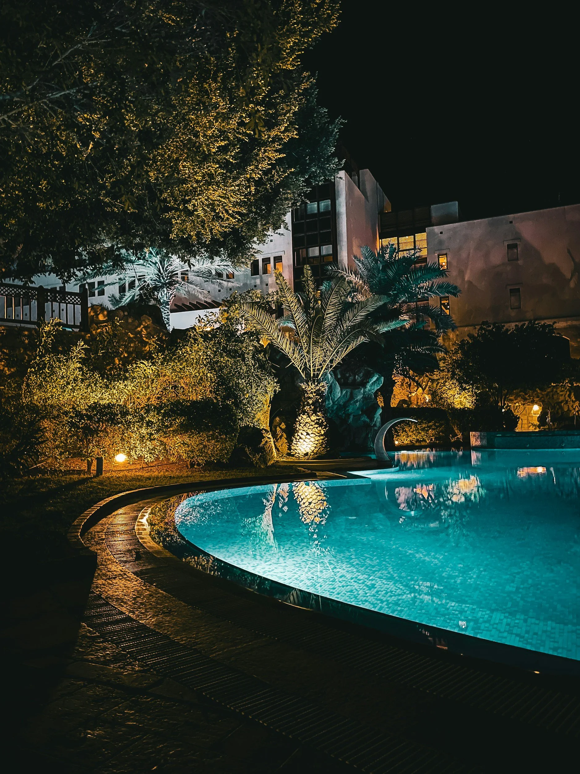 Nighttime scene of a lit swimming pool surrounded by lush plants and trees, with a modern building in the background.