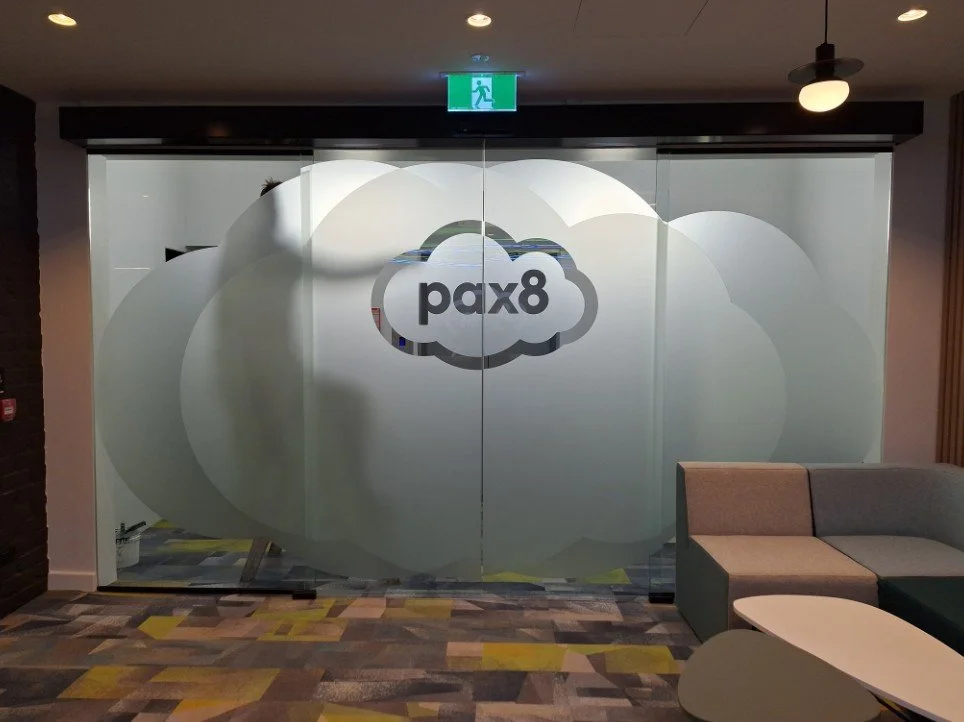 A glass office partition featuring a large, custom-frosted "Pax8" cloud logo for privacy and branding.