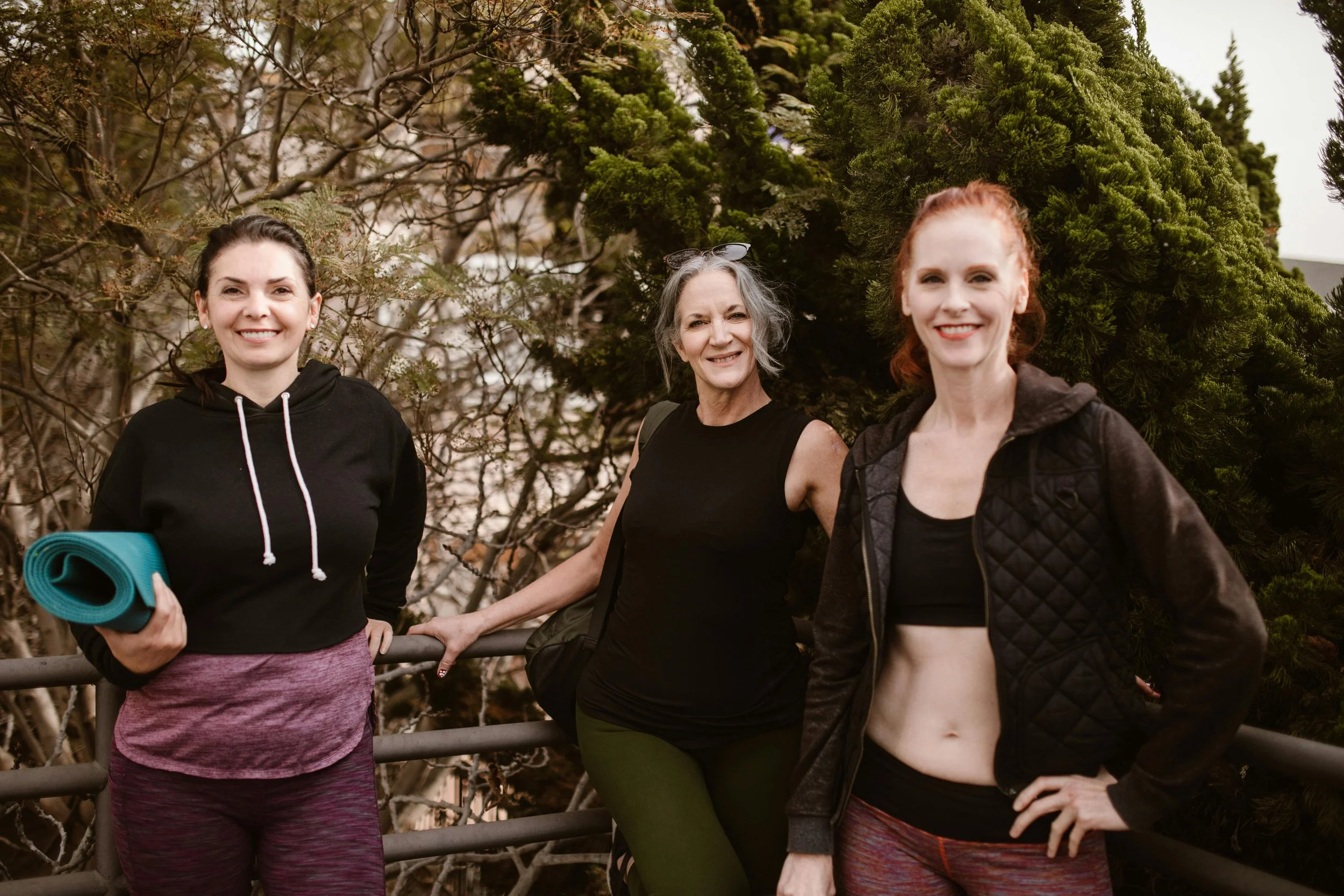 Three women pose outdoors near trees, dressed in workout clothes, smiling, with one holding a rolled-up yoga mat.