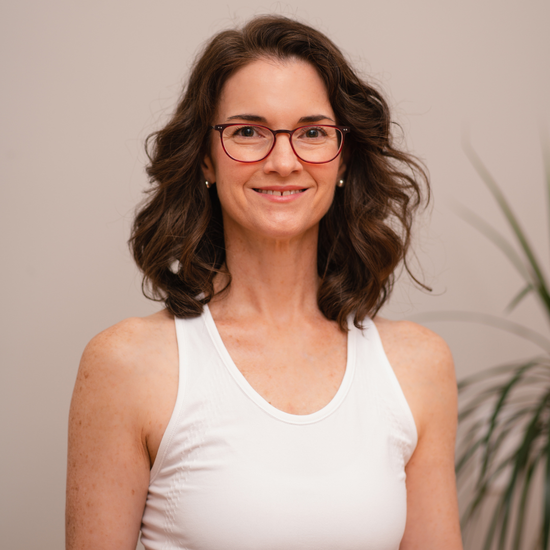 Cathy with shoulder-length curly brown hair, wearing red glasses, a white sleeveless top, and pearl earrings, standing in front of a plain light-colored wall with a plant in the background, smiling at the camera.