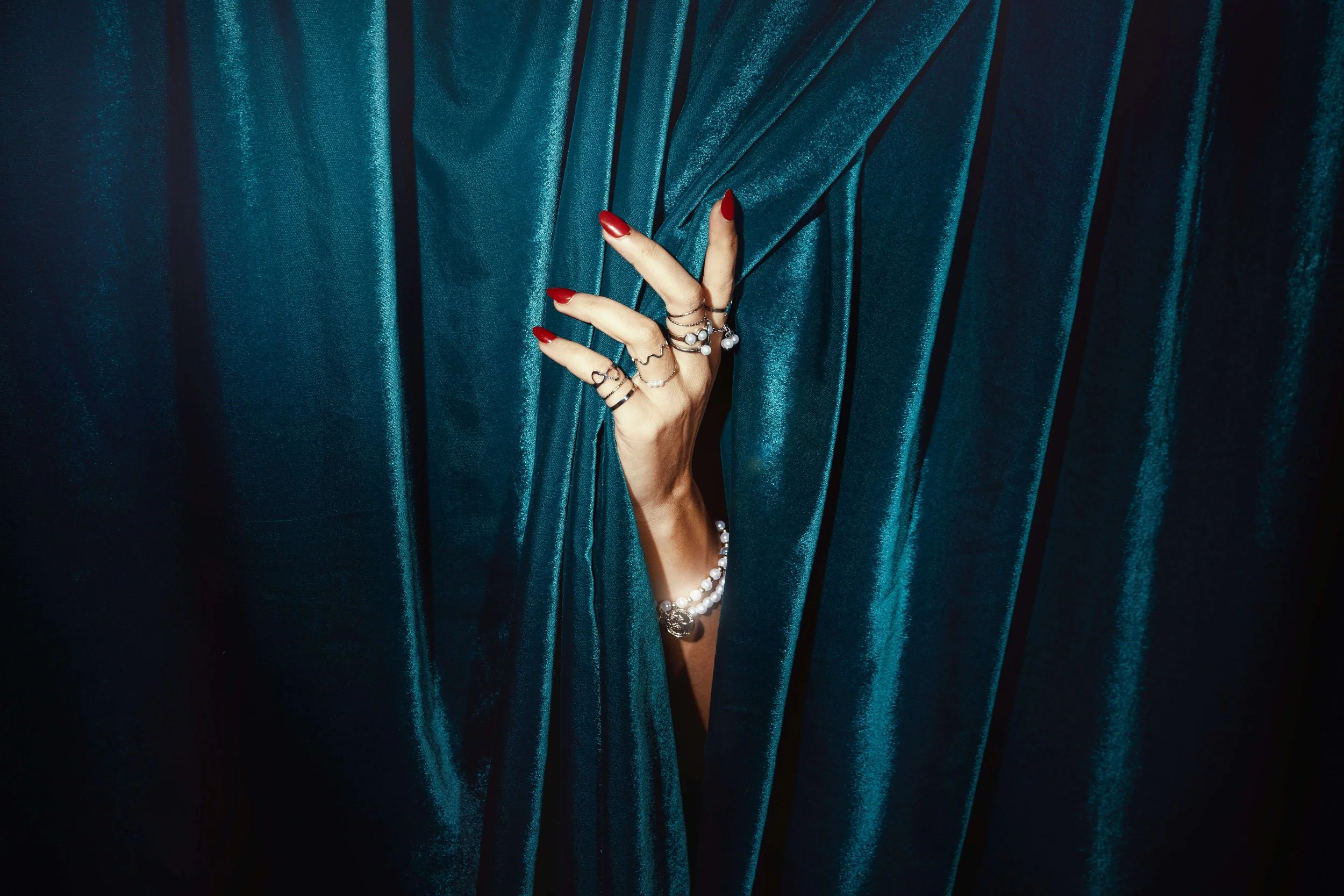 Hand with rings, bracelets and red nails Credit: Aditya Saxena - Unsplash poking out of a green velvet curtain 