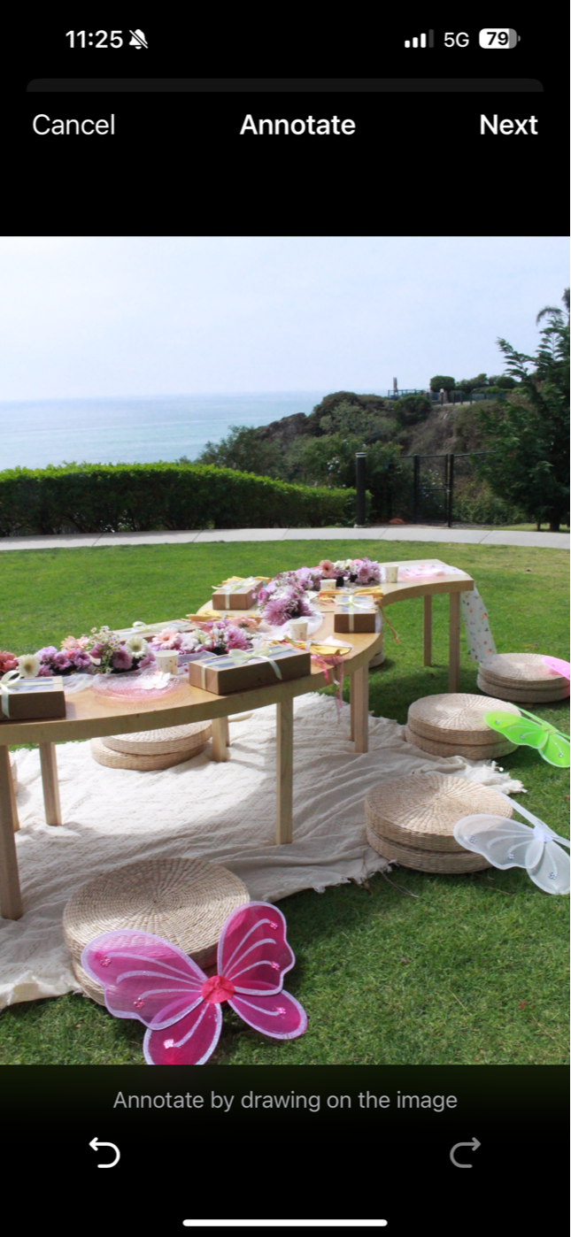 Empty beach with white chairs and tablecloth, ocean in the background, rocky cliff on the right, sunny sky, suggesting a seaside dining setup.