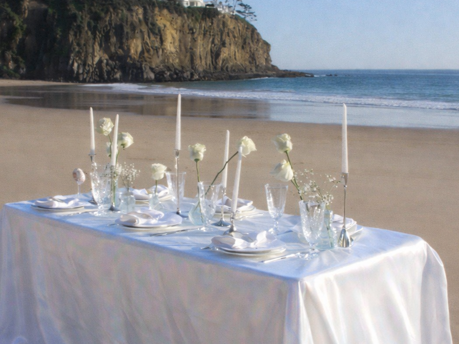 Elegant white table set for a special occasion on a beach, decorated with white roses, tall white candles, glassware, and napkins, overlooking the ocean and cliffs.