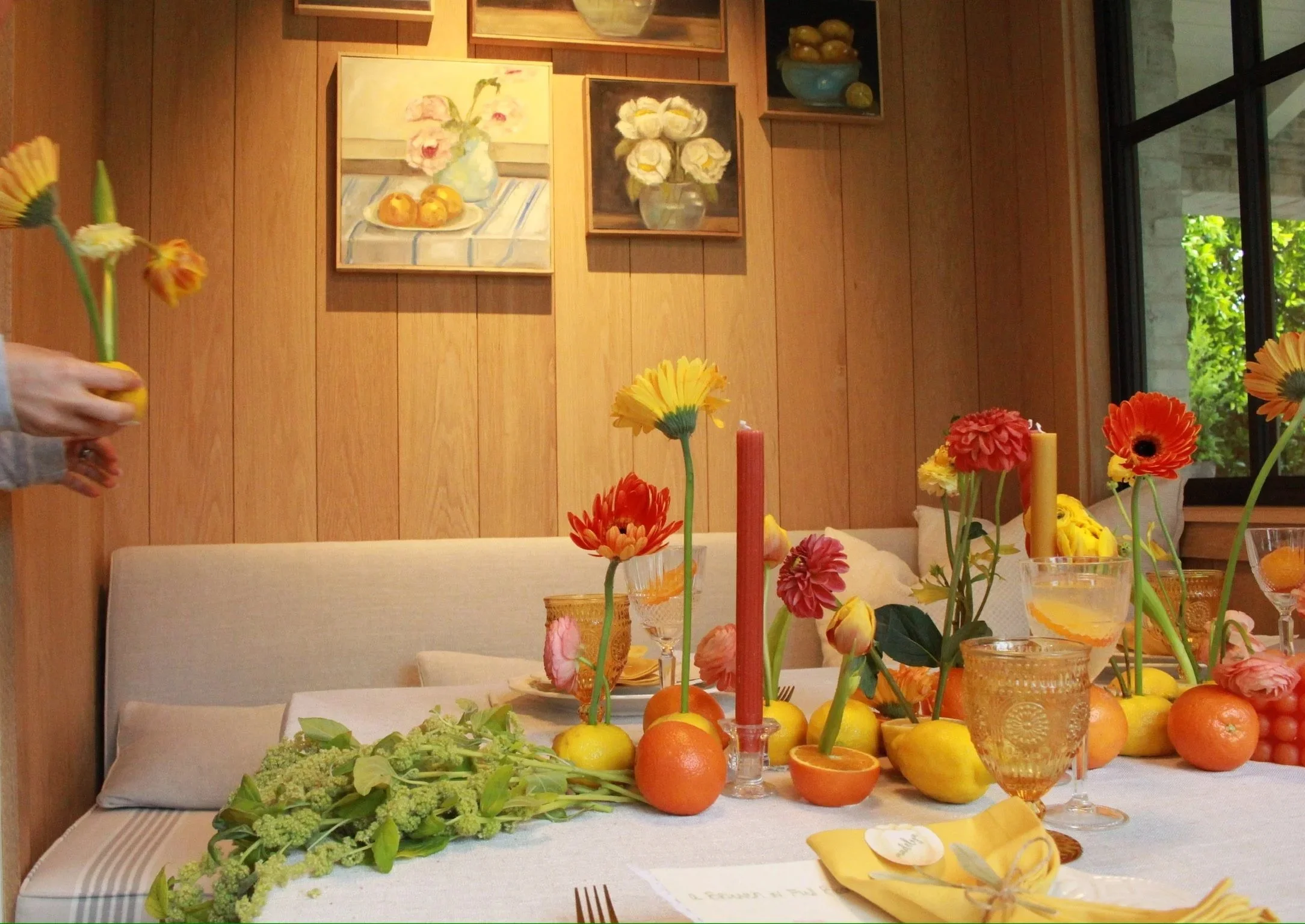 A dining table decorated with colorful flowers and fruits, with a wooden-paneled wall and framed artwork in the background.