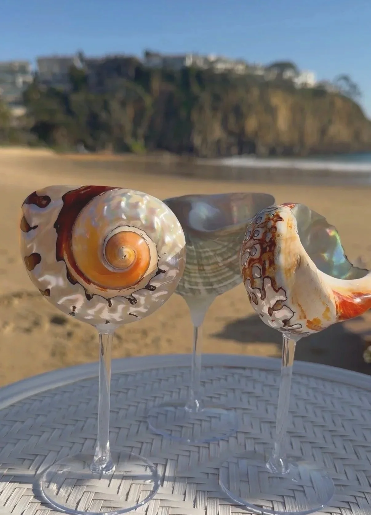 Three decorative glass shells on sticks are placed on a white circular table on a sandy beach, with cliffs and buildings in the background.
