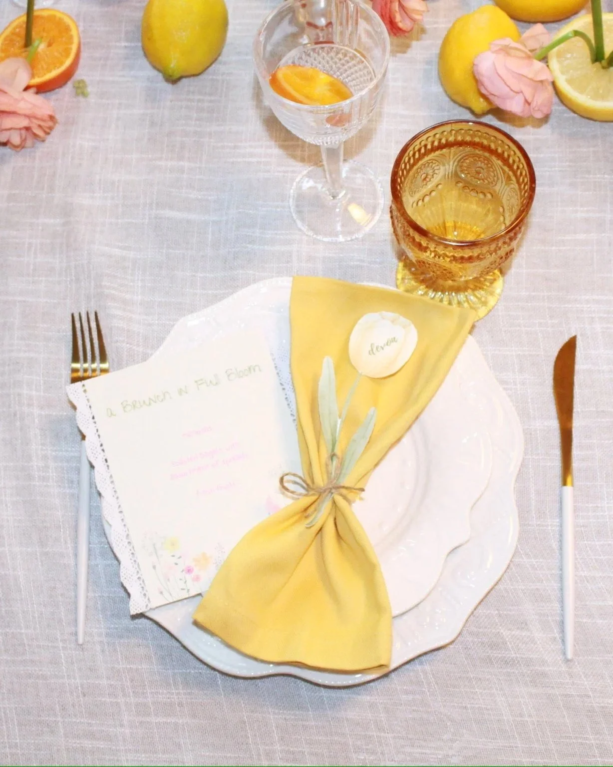 A table setting with a white plate, yellow napkin tied with a string containing a sprig and a small card, a fork and knife, two glasses with drinks and lemon slices, and some lemons, orange slices, and pink flowers on a beige tablecloth.