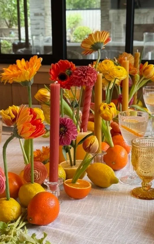 A colorful table centerpiece with assorted flowers in vases, surrounded by peels of oranges, lemons, and tangerines, with pink and yellow candles and glasses of drinks.