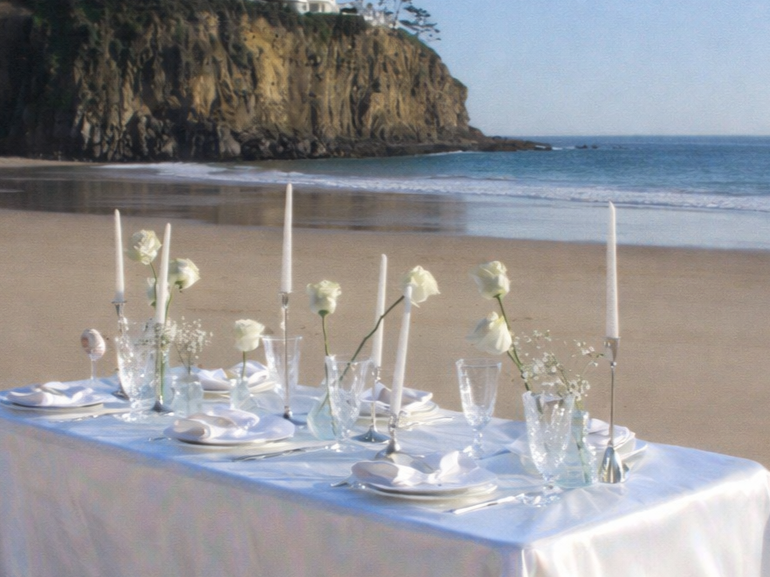 Elegant white table set for a special occasion on a beach, decorated with white roses, tall white candles, glassware, and napkins, overlooking the ocean and cliffs.