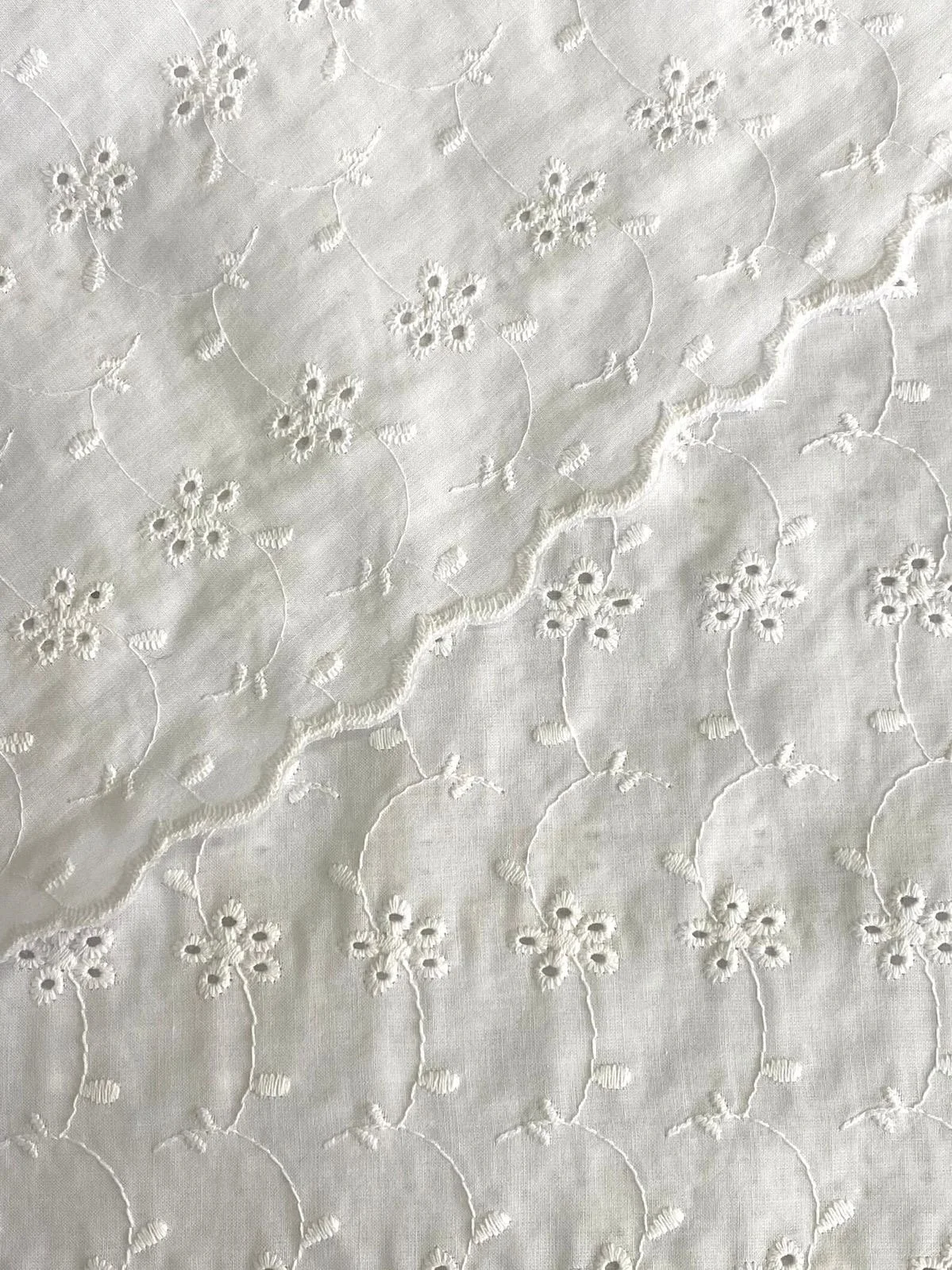 Close-up of white fabric with embroidered floral pattern, featuring small flowers connected by curving stems and scalloped edges.