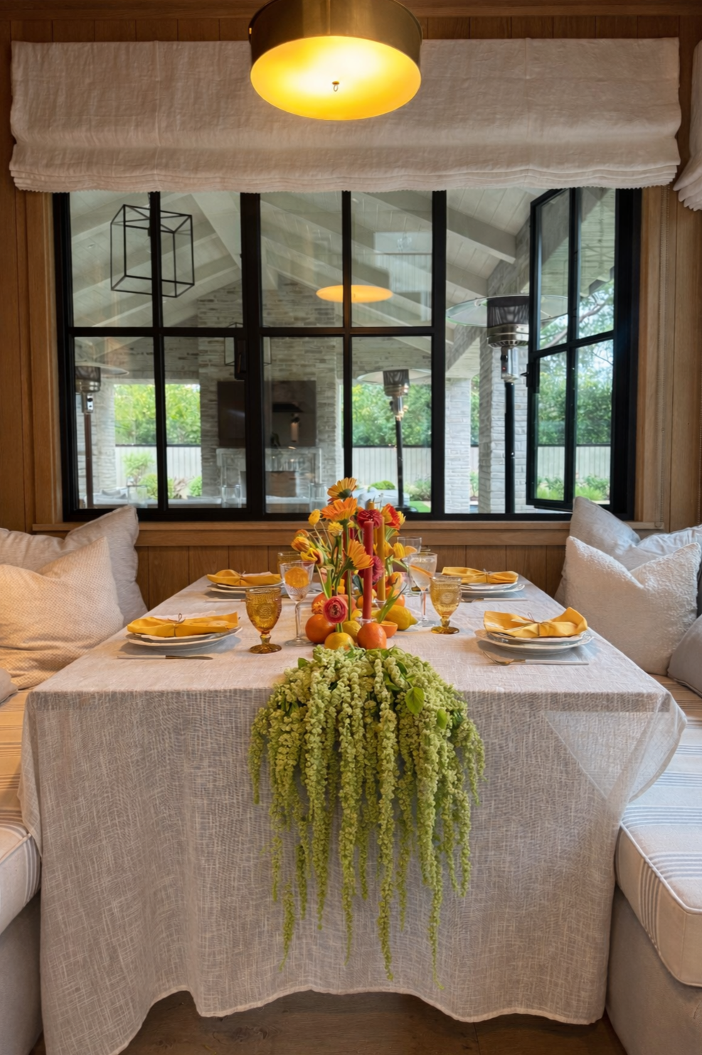 A dining table decorated for a meal with a floral centerpiece, yellow napkins, gold-rimmed glasses, and place settings. The table is set in a bright room with large windows looking out onto a patio with outdoor heaters and greenery.