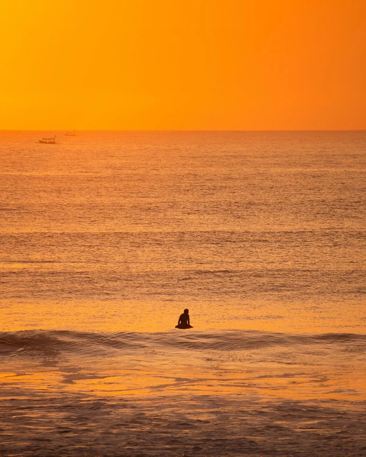 Person sitting on the beach near the water at sunset with orange and yellow sky, and boats visible in the distance.