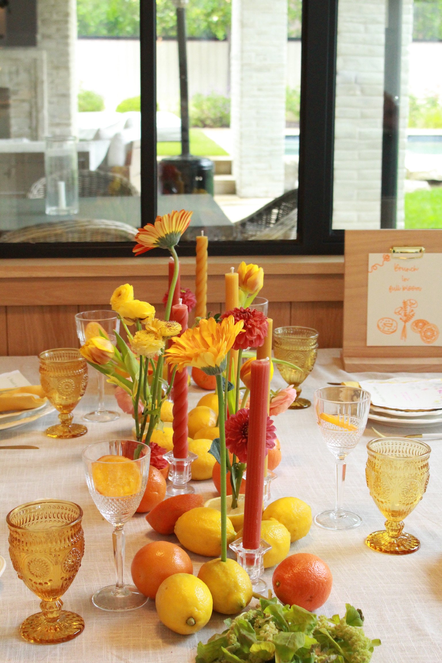 A table decorated with yellow, orange, and red flowers, lemons, grapefruits, and candles, set for a meal with glassware and plates, in a room with large windows revealing an outdoor patio area.