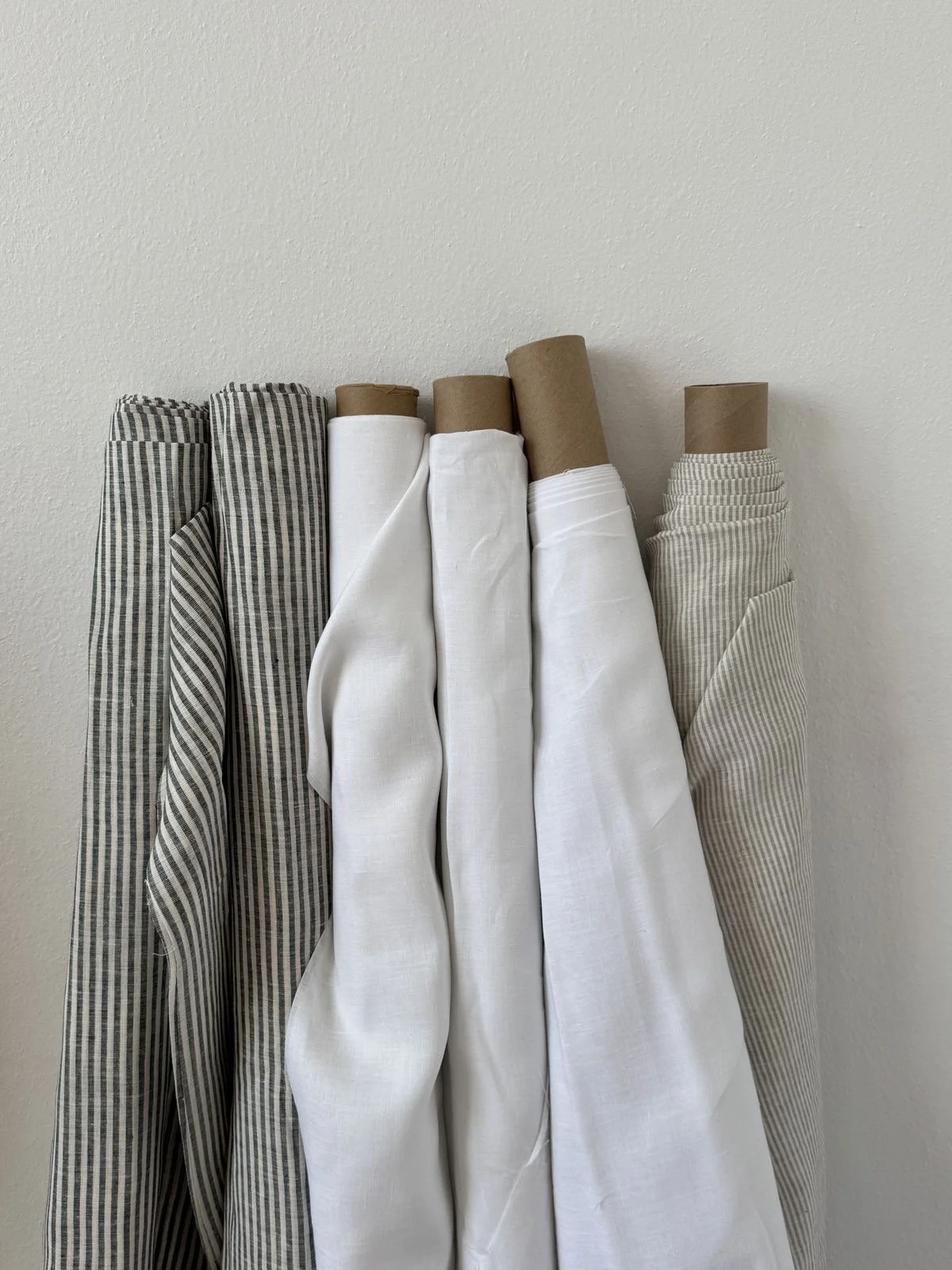 Assorted fabric bolts and rolls, mostly in neutral colors and patterns, leaning against a plain white wall.
