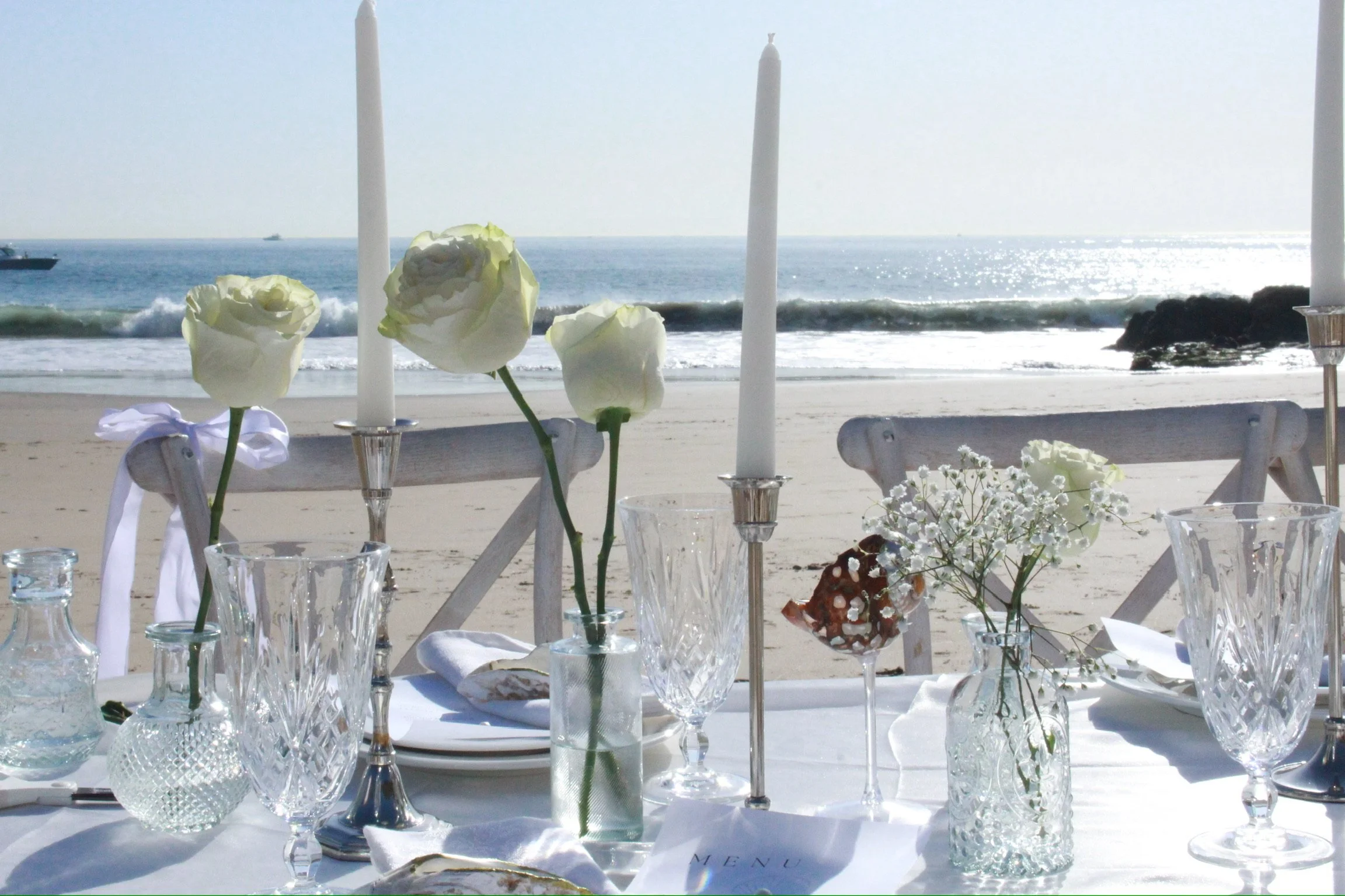 Beachside table set with white flowers, candles, and glassware, overlooking the ocean with boats in the distance.