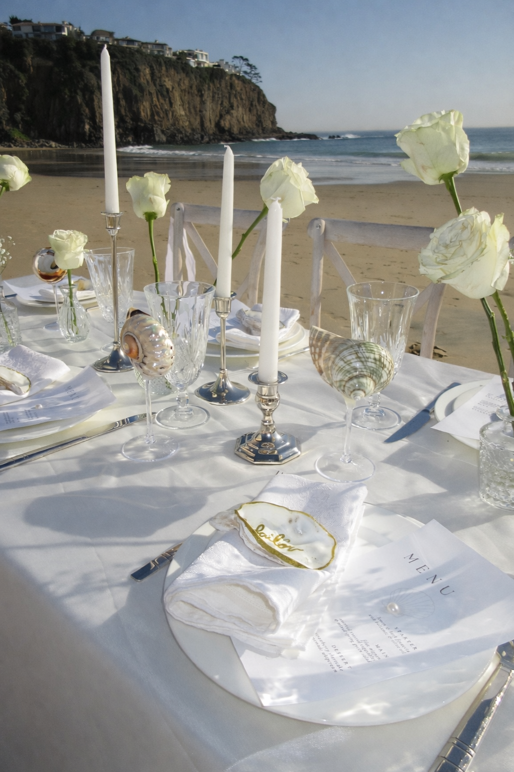 Elegant beachside table setting with white flowers, candles, shells, and a menu, overlooking a sandy beach and ocean cliffs.