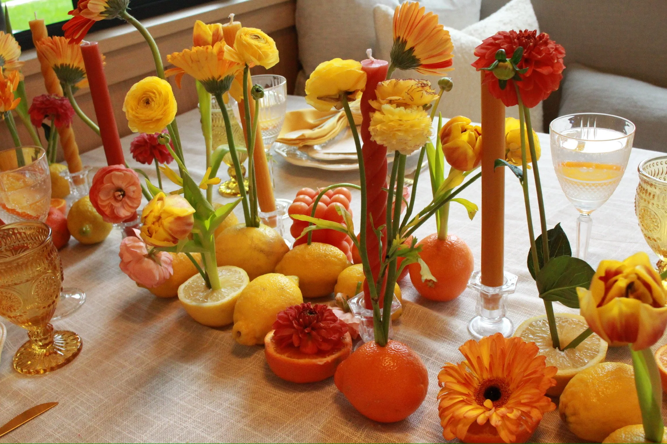 A table decorated with yellow, orange, and pink flowers, lemons, and oranges, with lit candles and glassware, set for a meal or celebration.