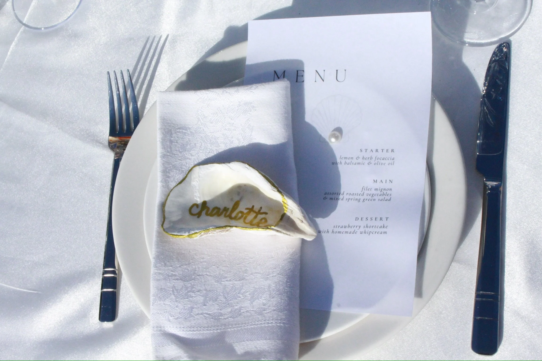 Elegant table setting with a white plate, silver fork and knife, a white napkin, a menu card, and a slice of cake with the name "Charlotte" written in gold on it, all on a white tablecloth.