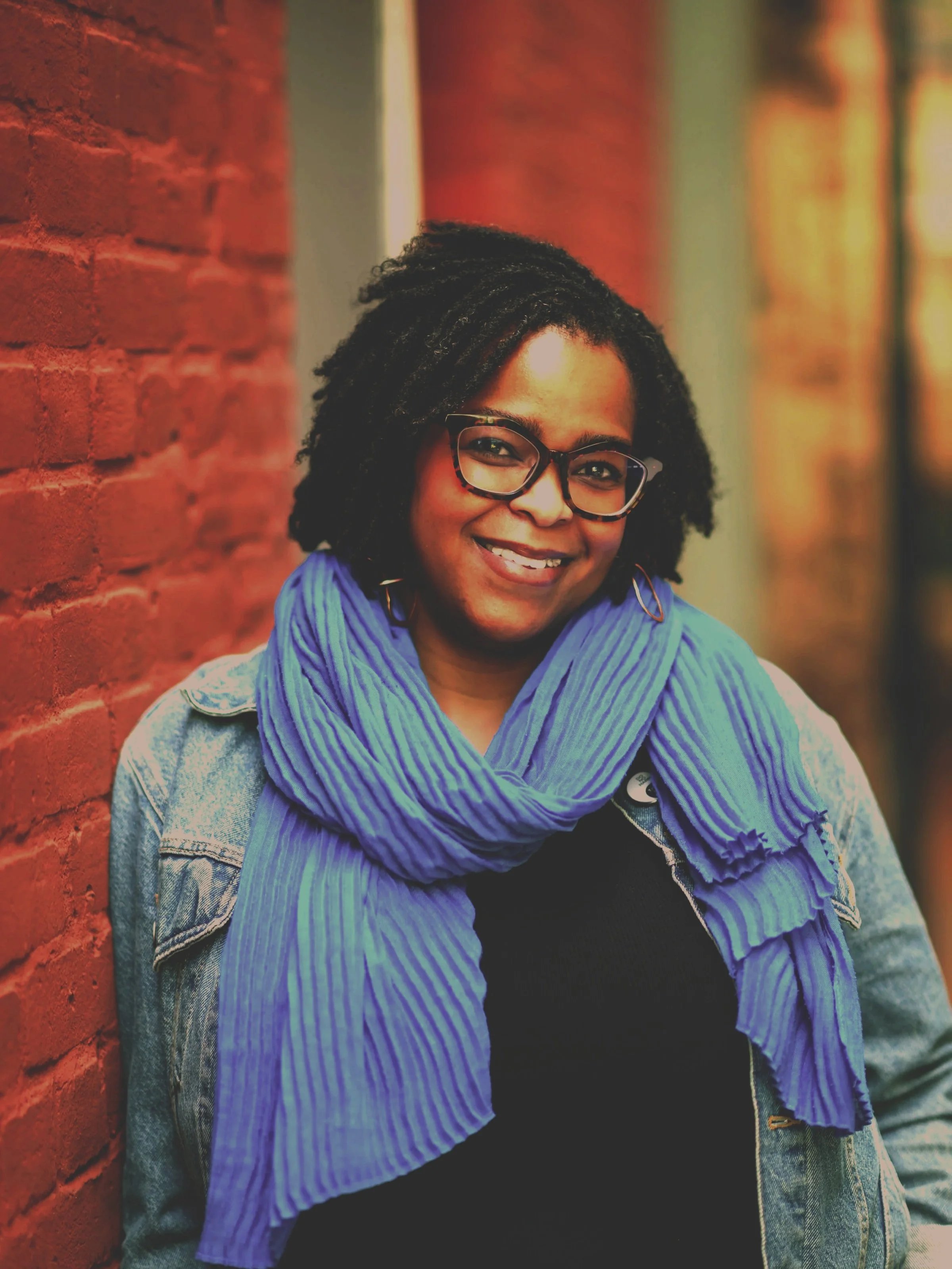 Novera Alim King. Developmental editor, writer, teacher. Black woman with locs and glasses leans against brick wall.