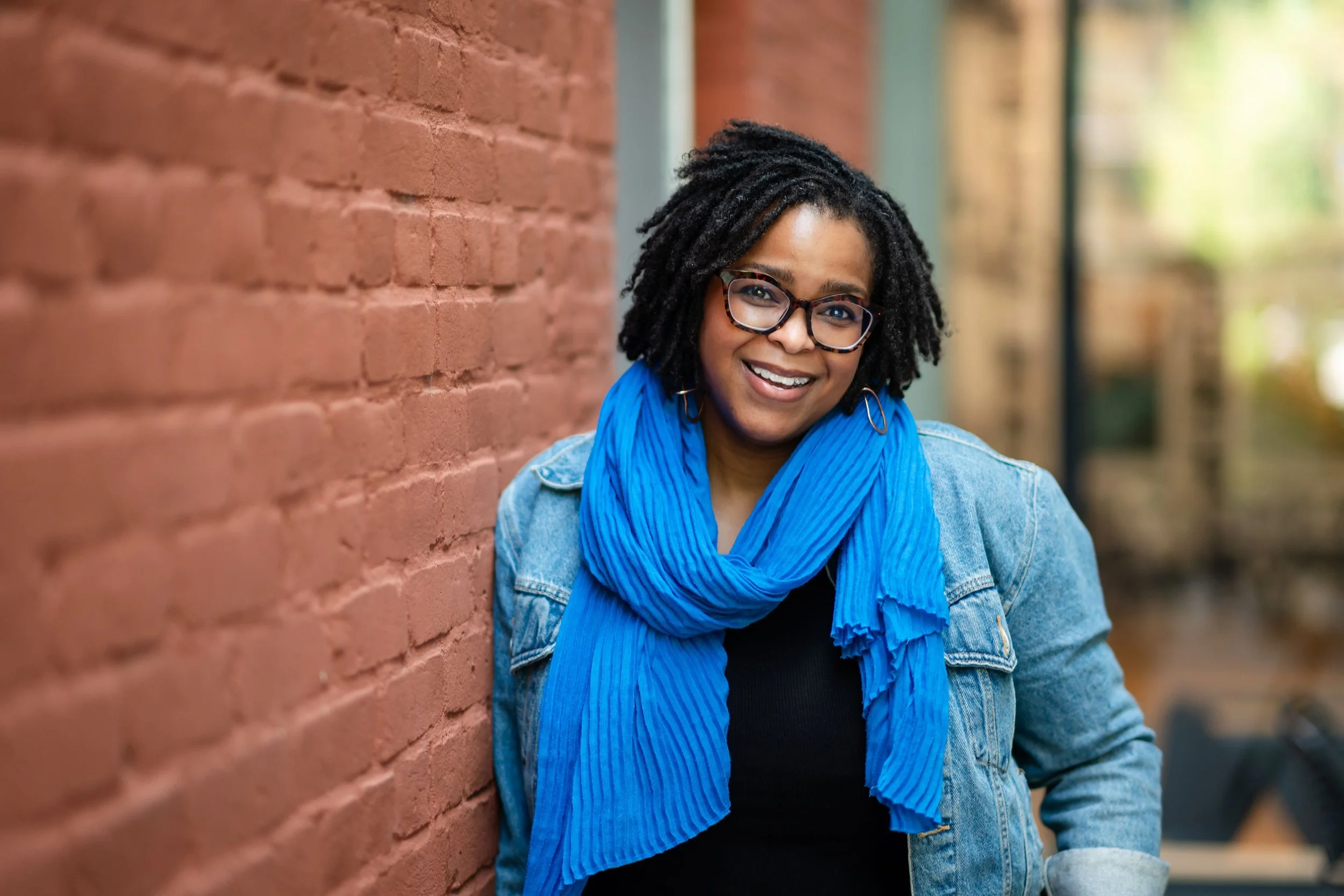 Novera Alim King, writer, editor, and story consultant. She's a Black woman with locs, wearing eyeglasses, a smile, and a bright blue scarf, and leaning against a brick wall.