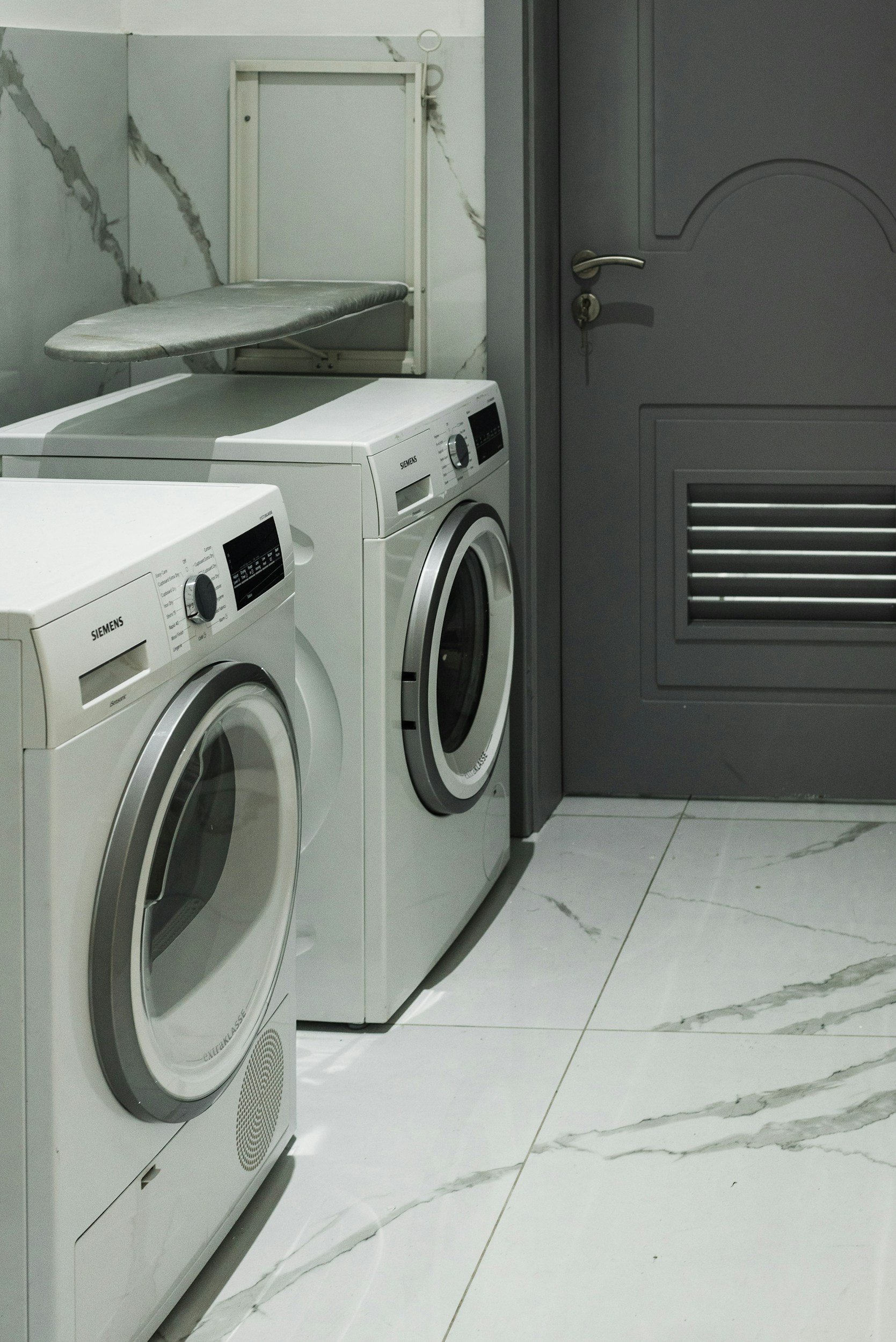 A laundry room with two front-loading washing machines, a gray door, and an ironing board on the wall.