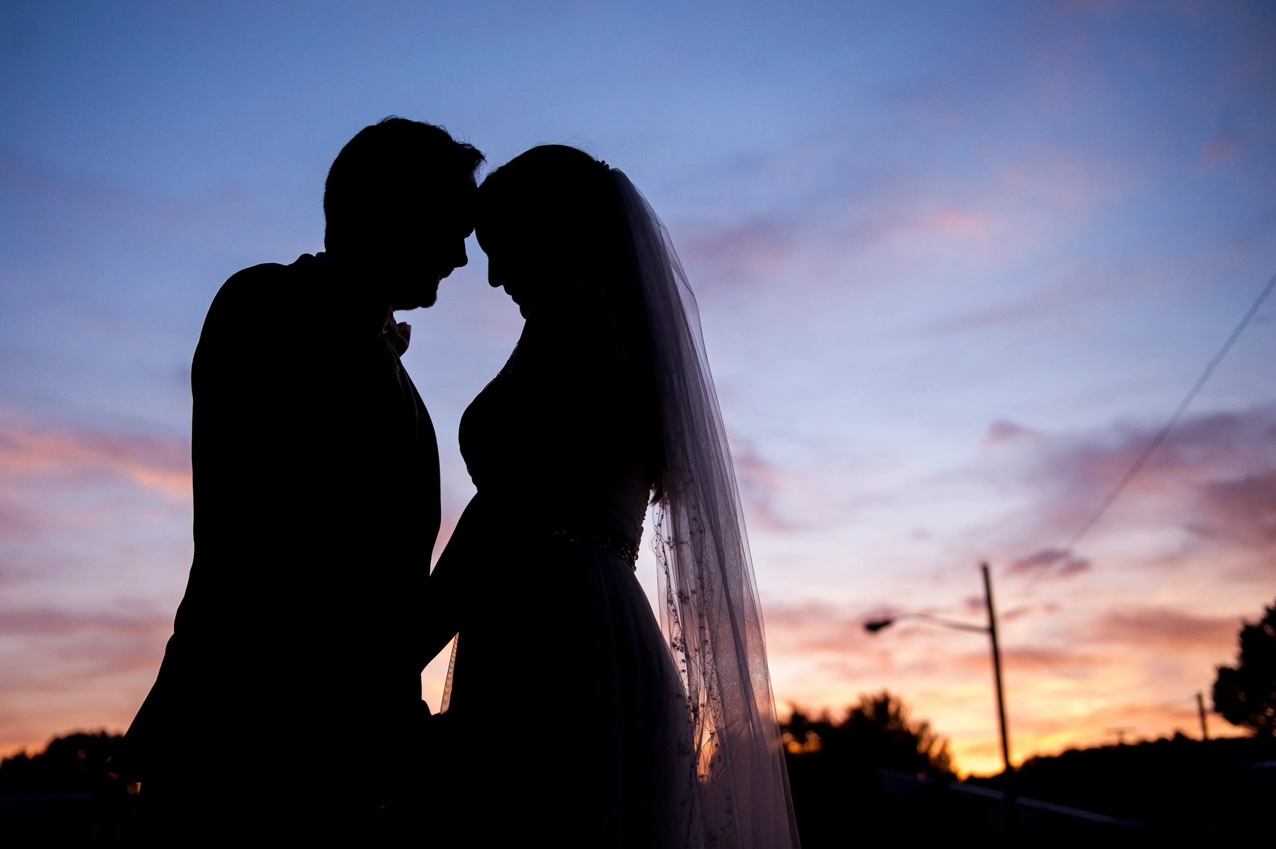 Silhouette of a couple, possibly newlyweds, standing close together with foreheads touching during sunset, with the sky having pink and purple hues.