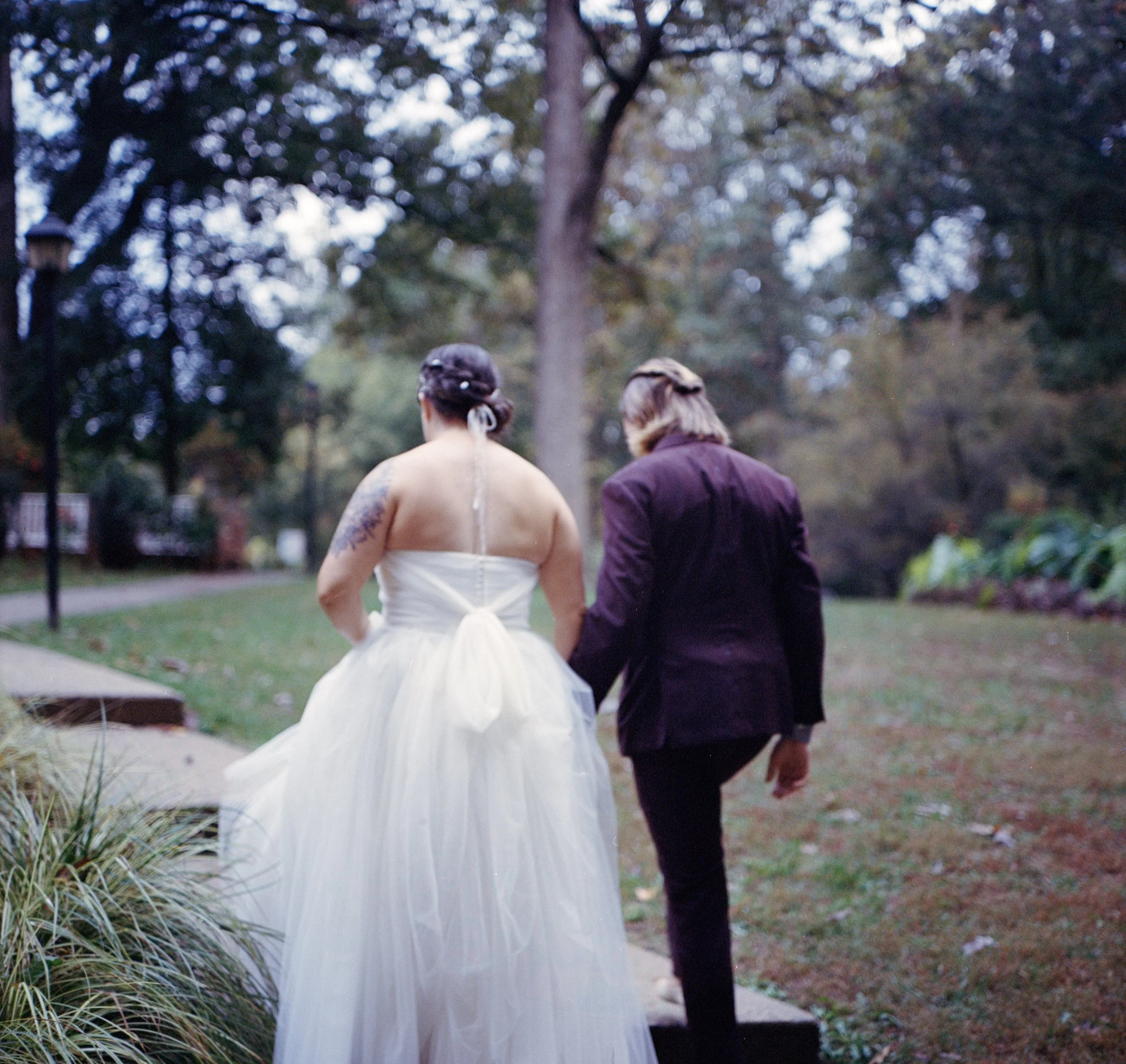 A couple dressed in wedding attire walking outside in a park, surrounded by trees and greenery.