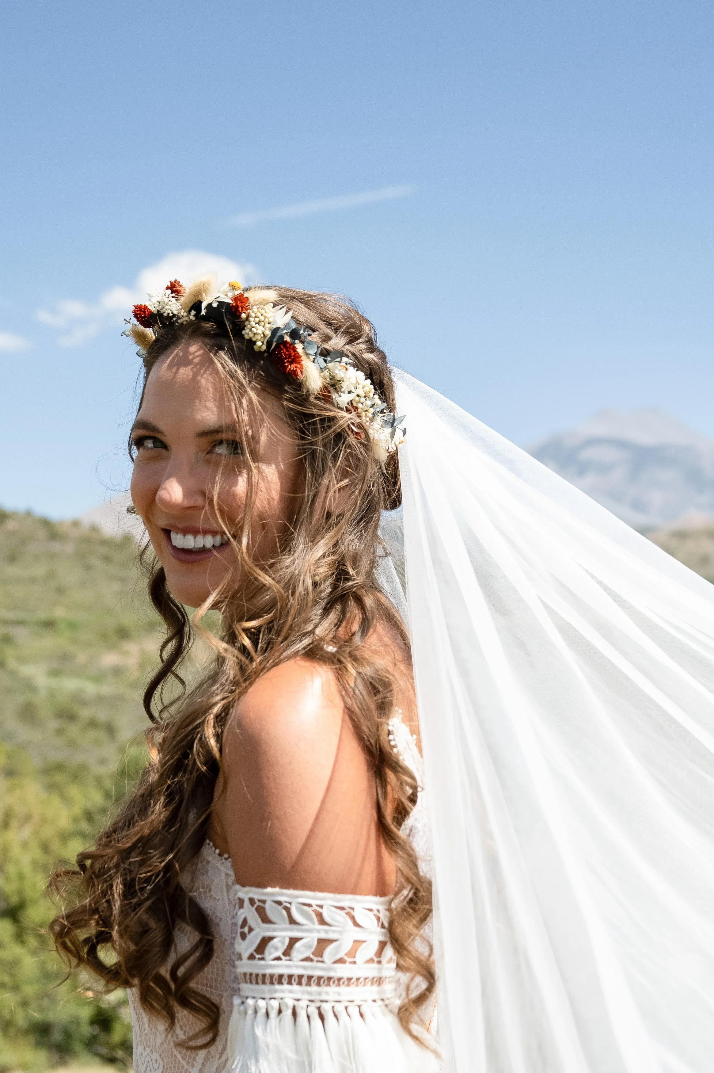 A woman in a wedding dress with a floral crown and veil outdoors on a sunny day, with mountains and blue sky in the background.