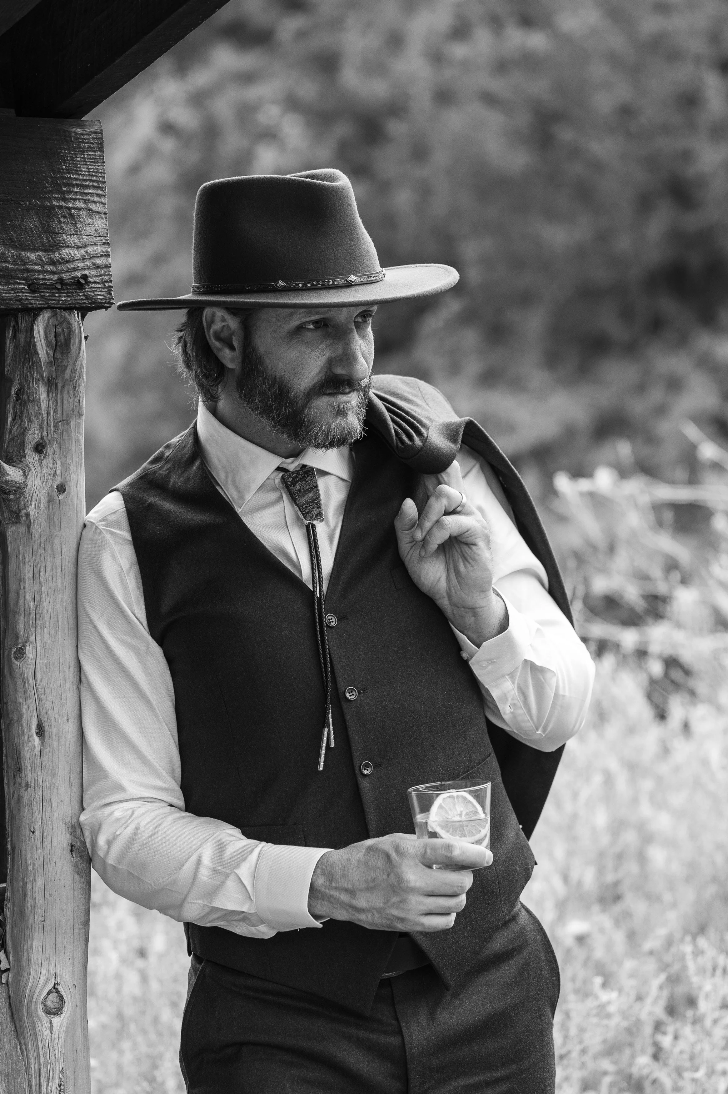 A man dressed in western-style clothing, wearing a wide-brimmed hat, vest, and shirt, leaning against a wooden post outdoors. He is holding a glass with a lemon slice and appears relaxed, with a jacket draped over his shoulder.