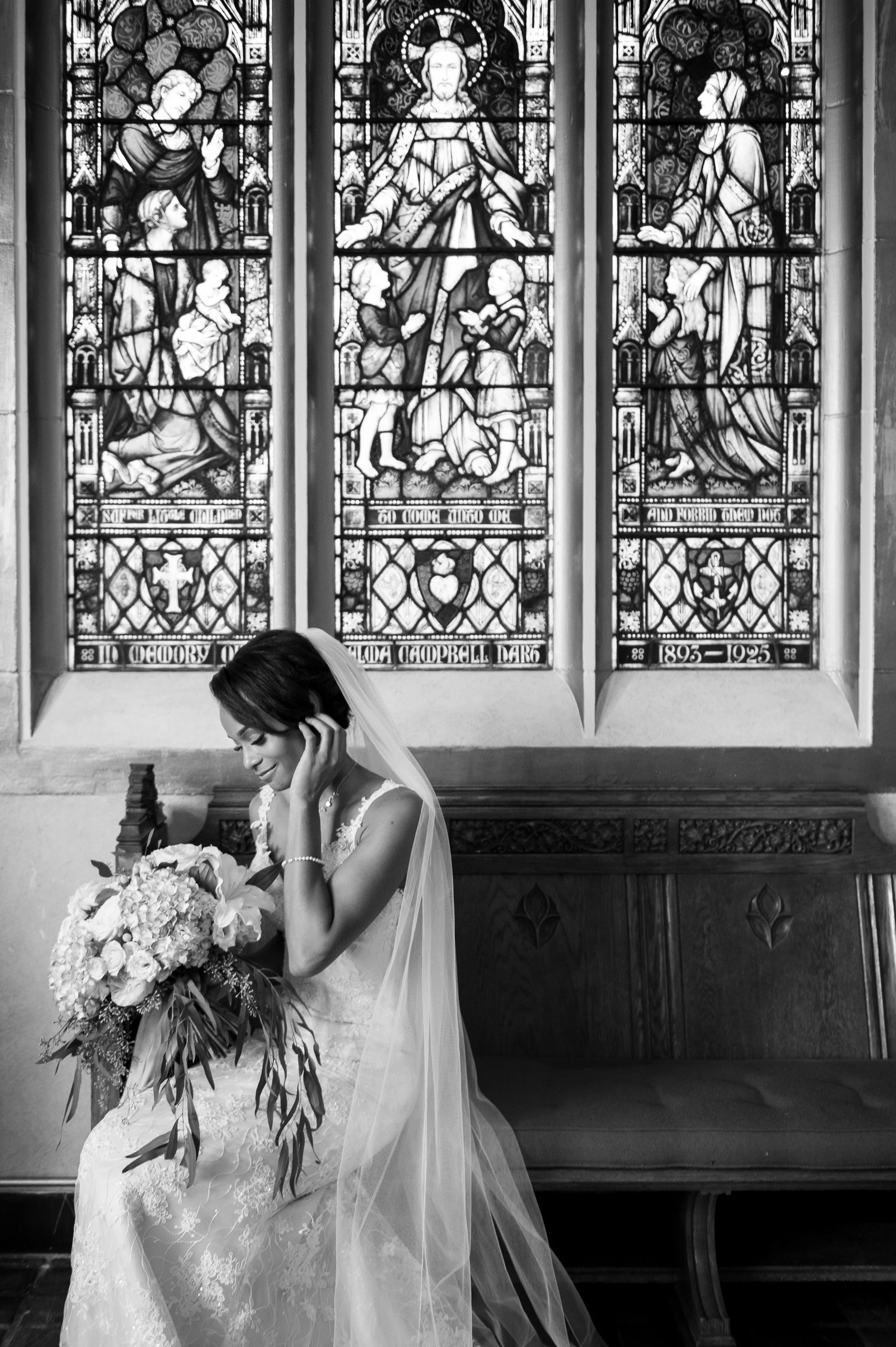 A bride sitting on a wooden bench in a church, holding a large bouquet of flowers, wearing a lace wedding dress and veil, with stained glass windows in the background.