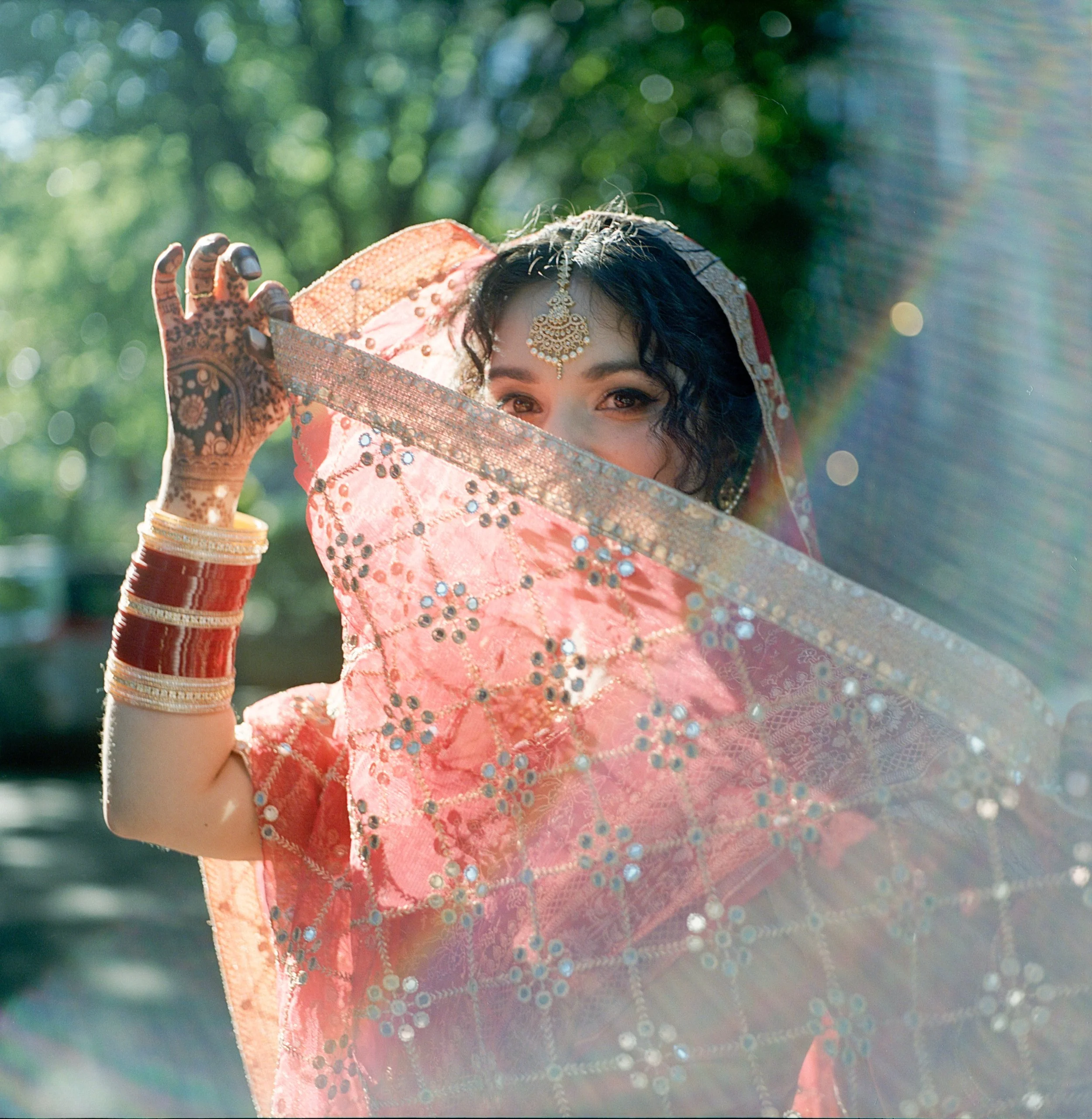 A woman dressed in traditional Indian attire, holding a pink embroidered veil in front of her face with one eye visible, outdoors with sunlight filtering through trees.
