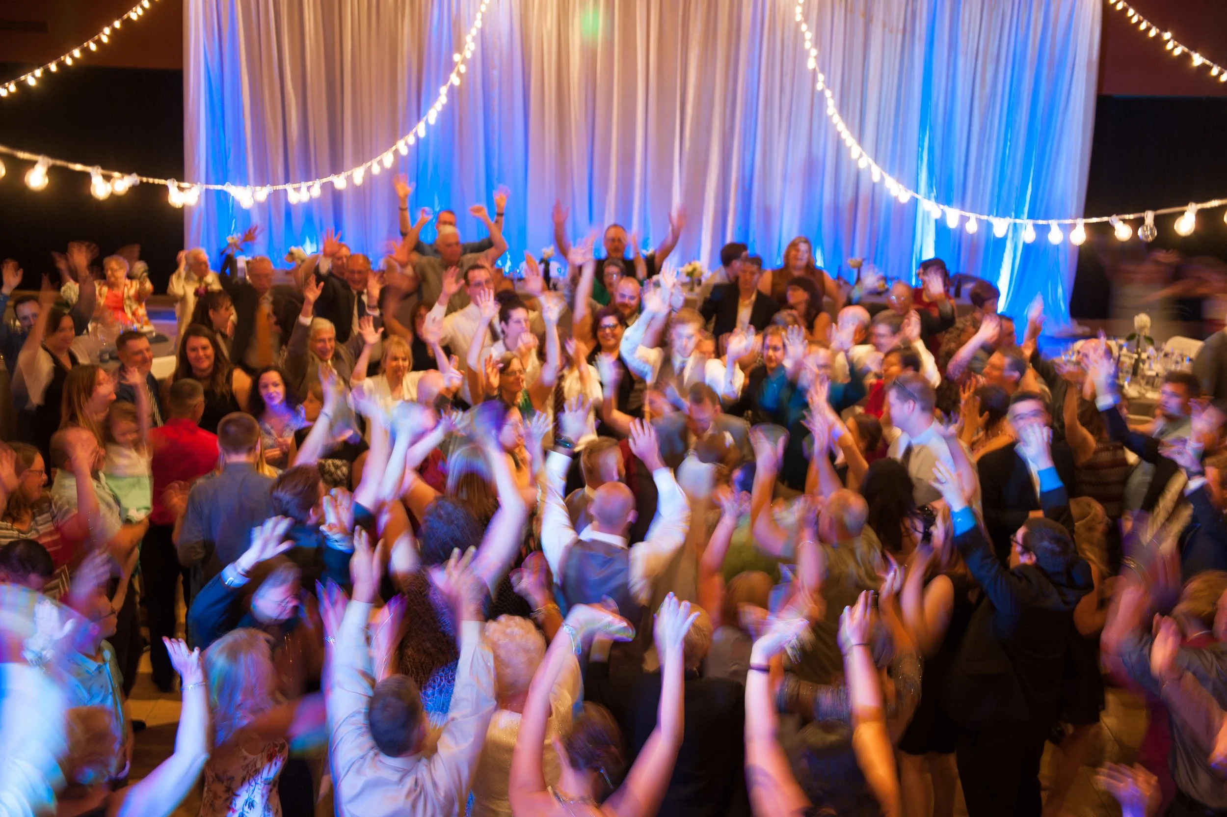 A lively party or celebration with a large group of people dancing and raising their hands under string lights in a decorated indoor venue with blue lighting and draped fabric backdrop.