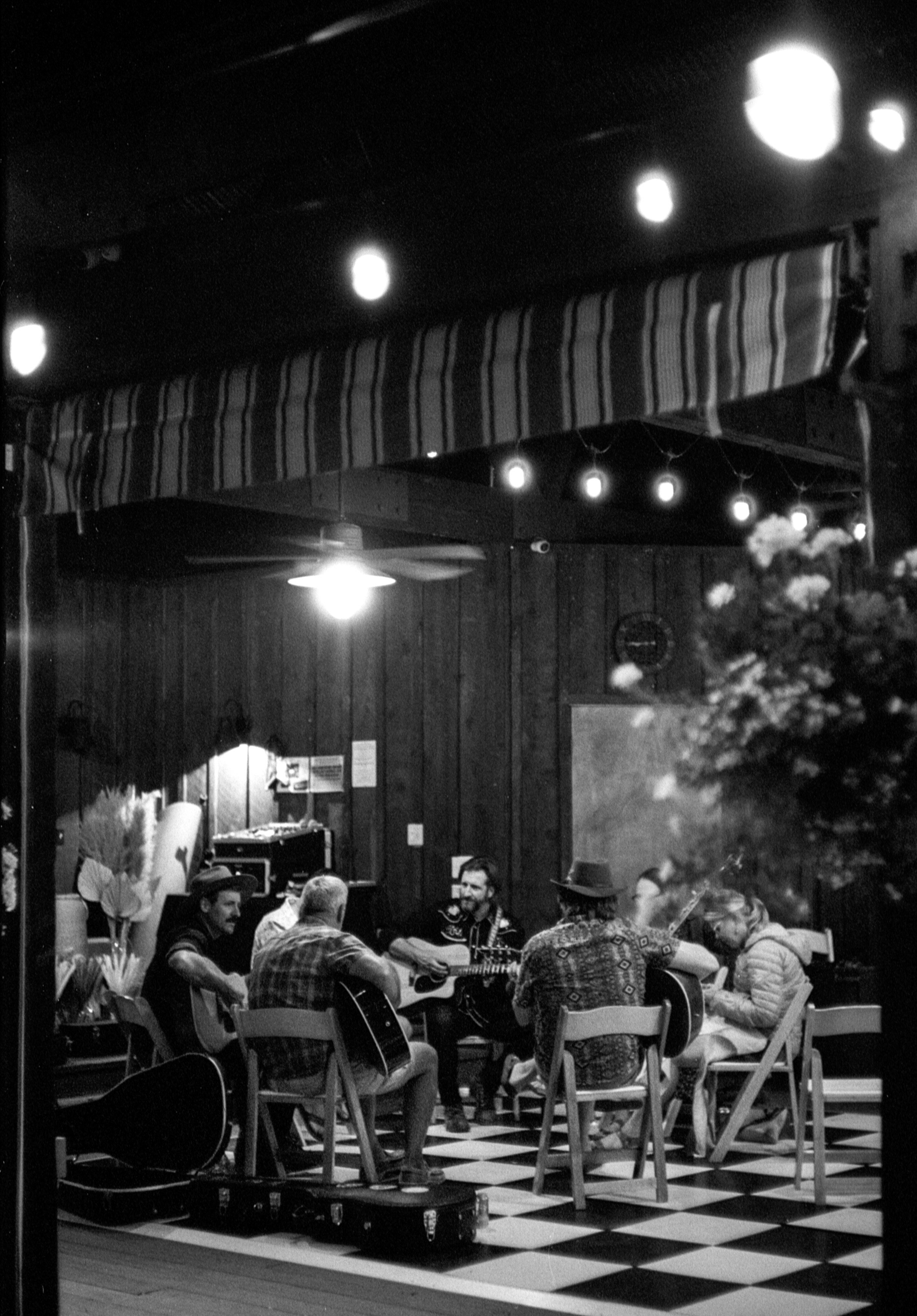 A group of musicians playing guitars in a dimly lit, cozy indoor setting with a checkered floor, string lights, and wooden walls.