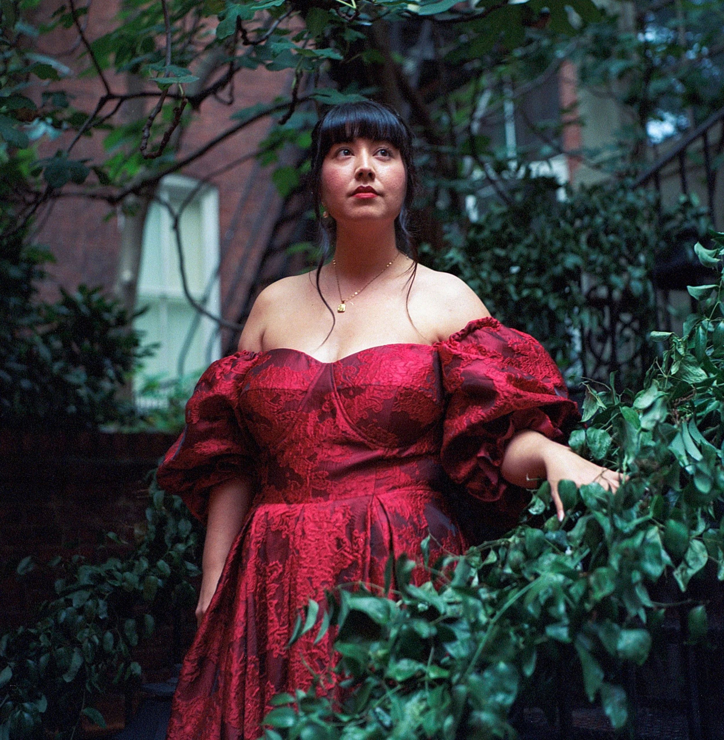 A woman in a red, off-shoulder dress standing among green foliage outdoors.