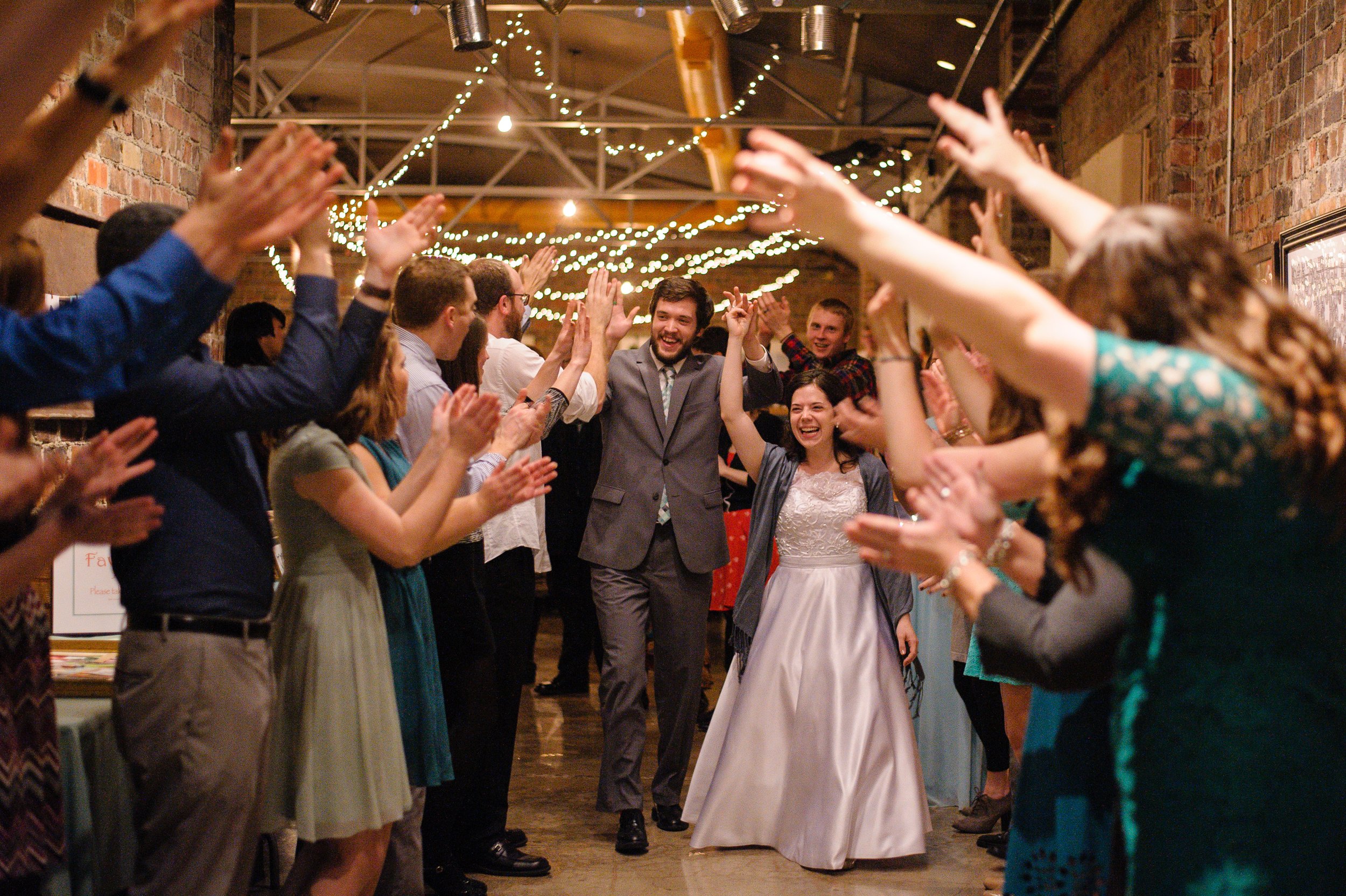A group of people celebrating a wedding reception, with a bride and groom walking through a tunnel of guests with raised hands, in a decorated venue with string lights on the ceiling.