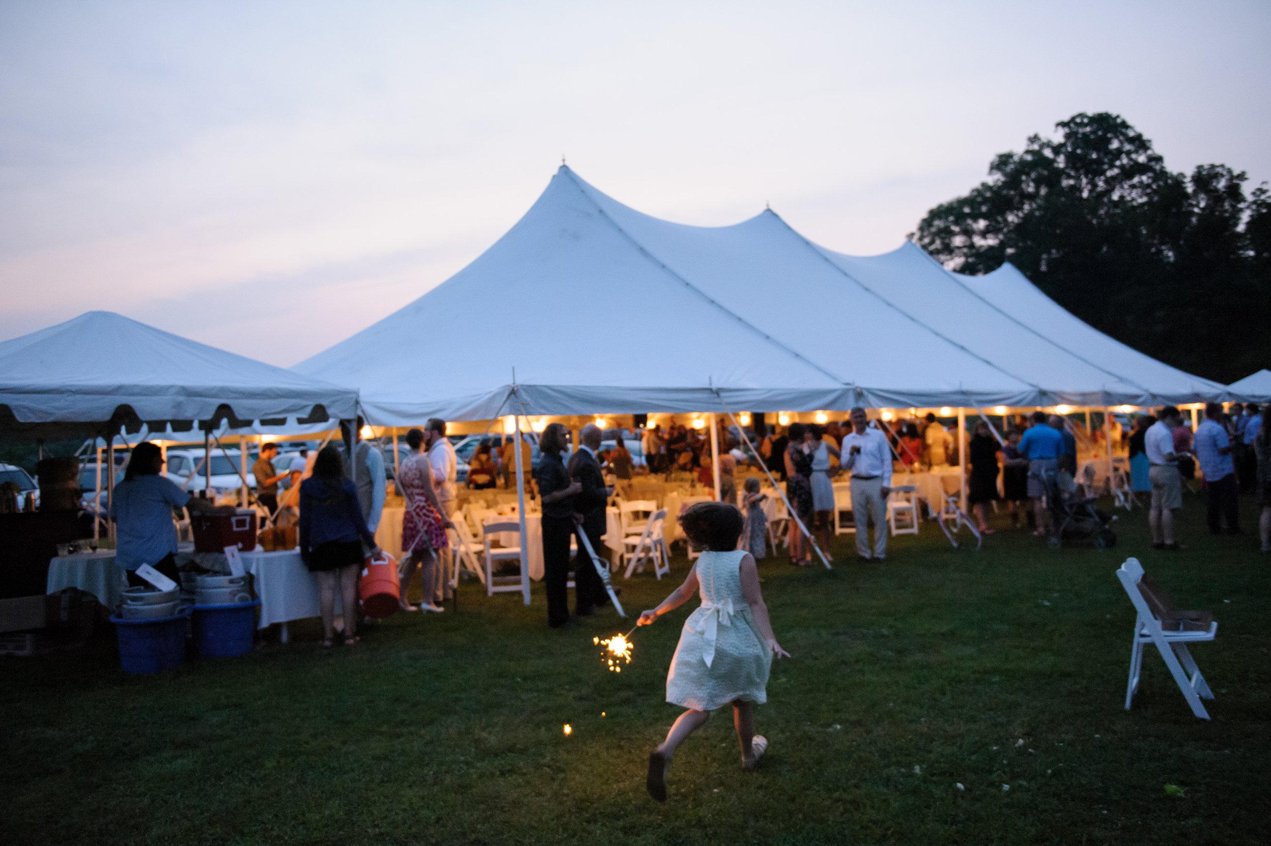 A girl runs with a sparkler in front of a large outdoor tent at dusk, with people gathered around and a tree in the background.