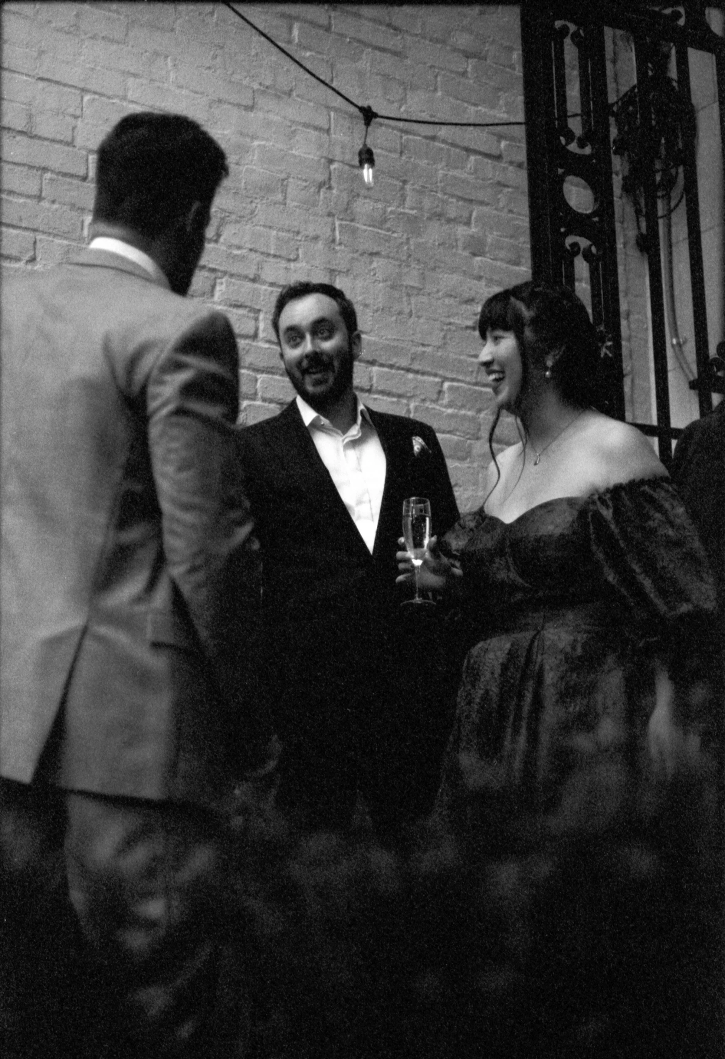 Three people talking at a social event, one woman holding a champagne glass, smiling, with two men, one with a beard, all dressed in formal attire, with a brick wall background.