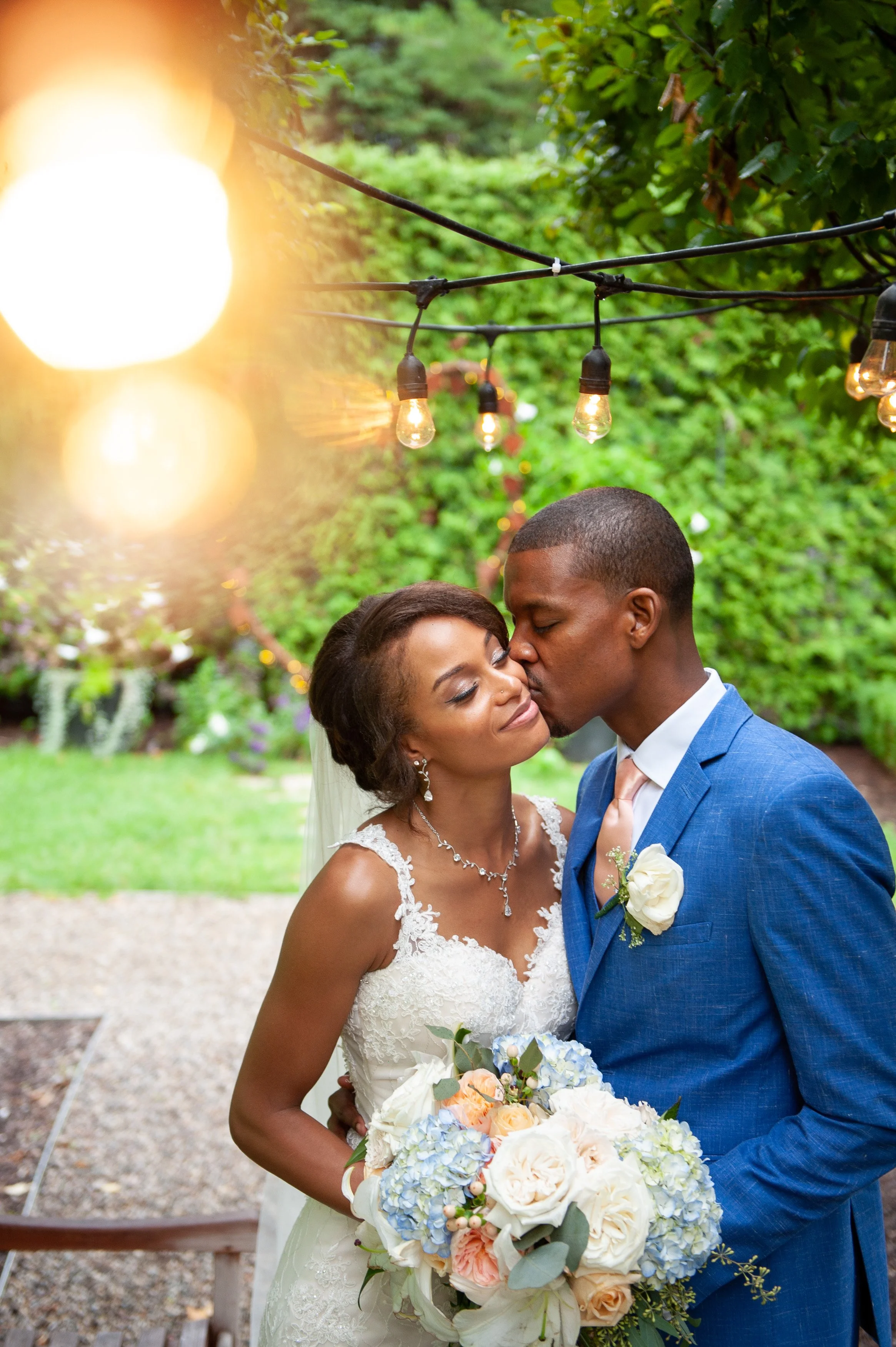 A newlywed couple sharing a kiss outdoors. The bride is holding a bouquet of flowers, and they are under string lights.