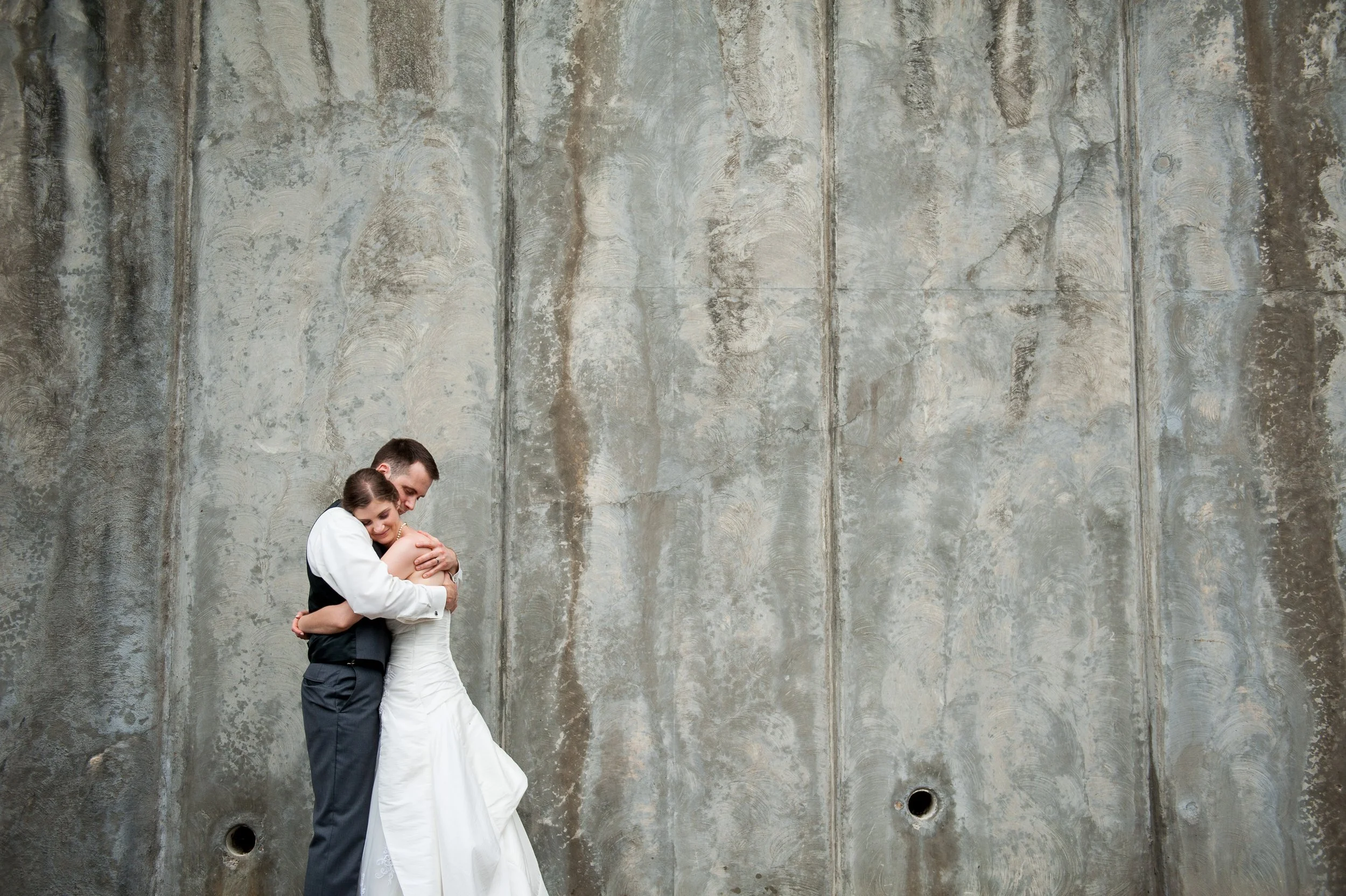 A newlywed couple hugging in front of a large concrete wall.