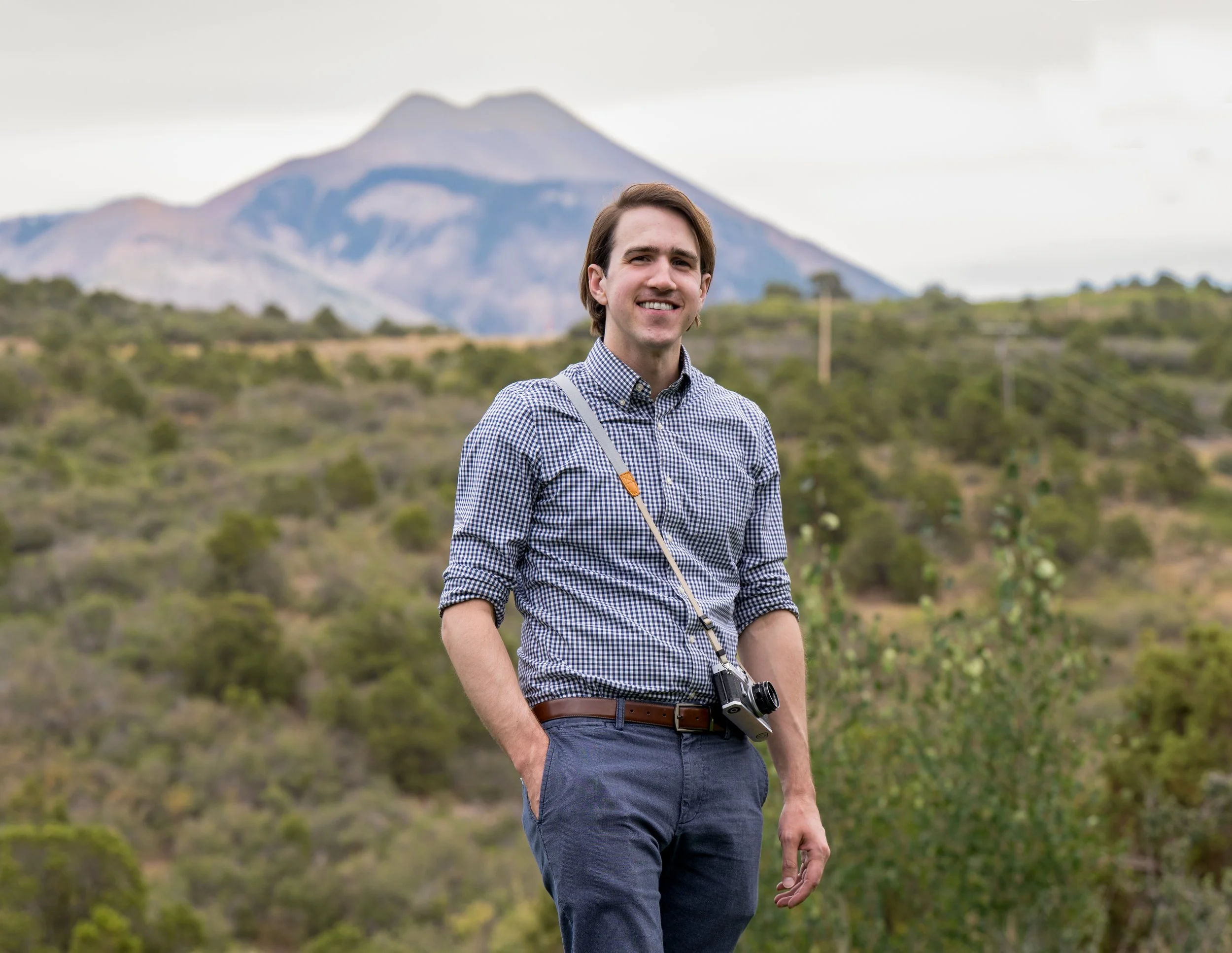 Greg Bodwell wearing a checkered shirt and gray pants, standing outdoors in a hilly area with mountains in the background, carrying a camera around his neck and smiling.