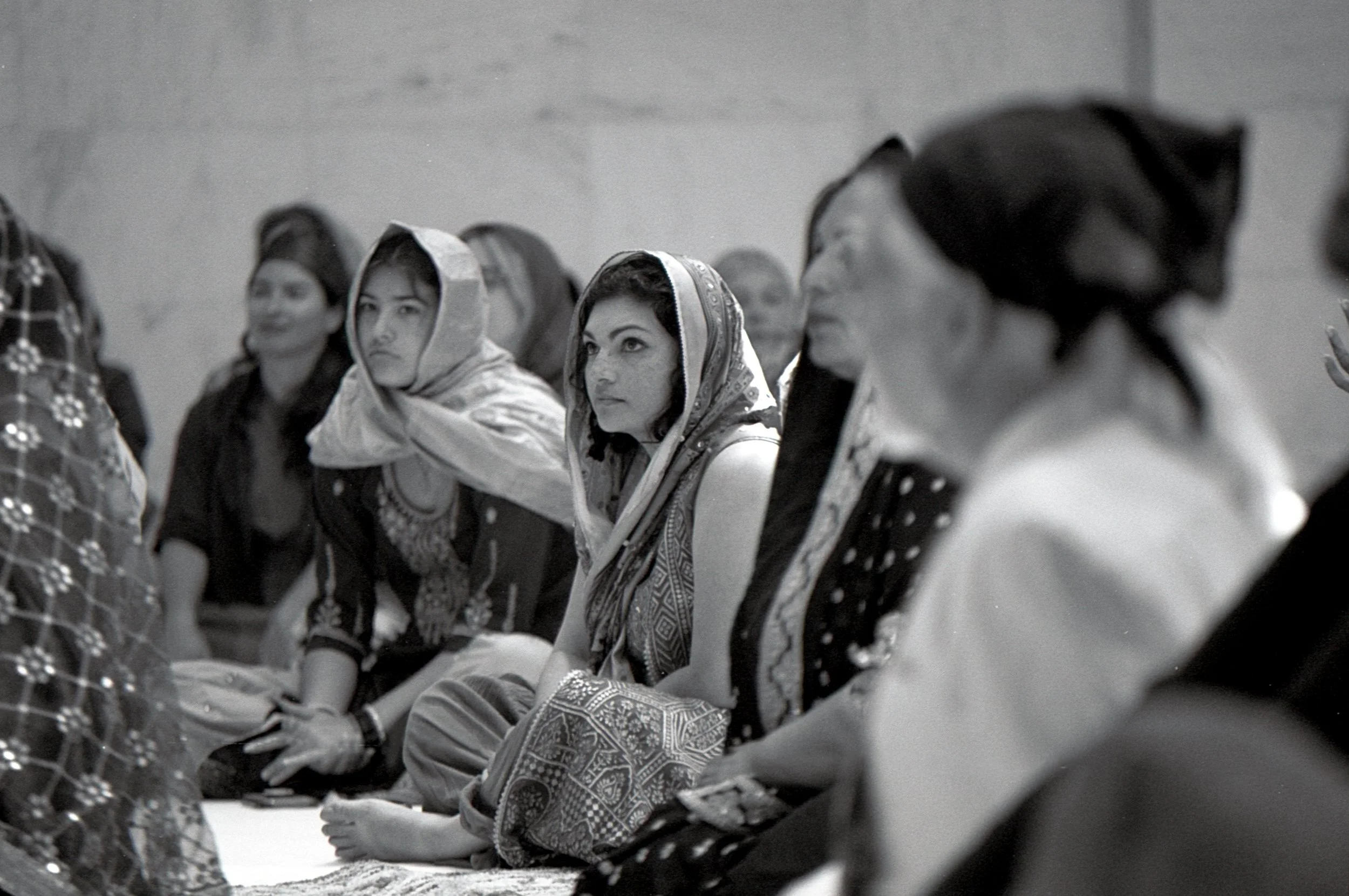 Group of women sitting on the floor, dressed in traditional South Asian attire, paying attention during a cultural or religious gathering, with some wearing headscarves.