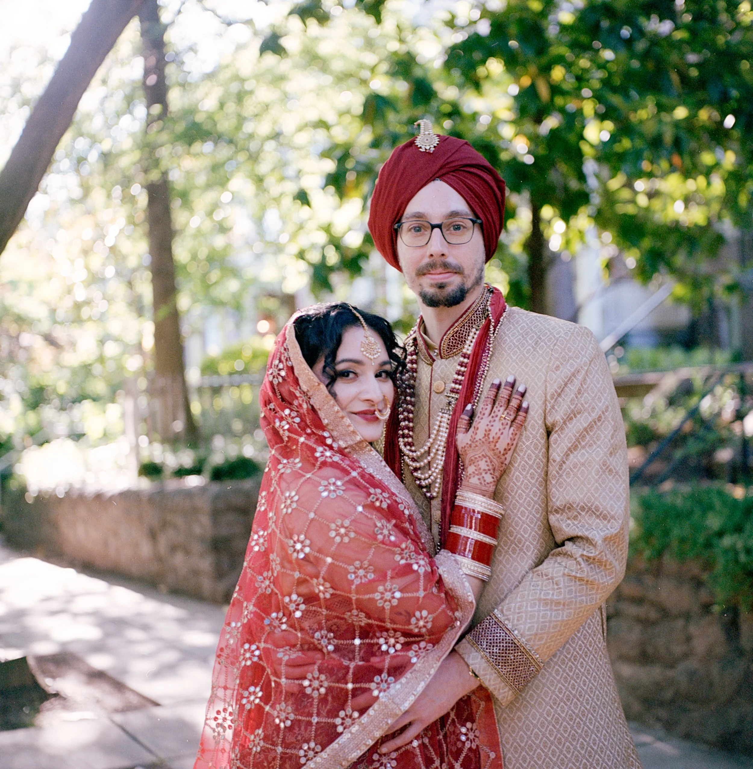 A couple dressed in traditional Indian wedding attire standing outdoors. The woman is wearing a red sari with intricate embroidery and jewelry, including bangles and a nose ring. The man is wearing a beige sherwani with a red turban decorated with a 