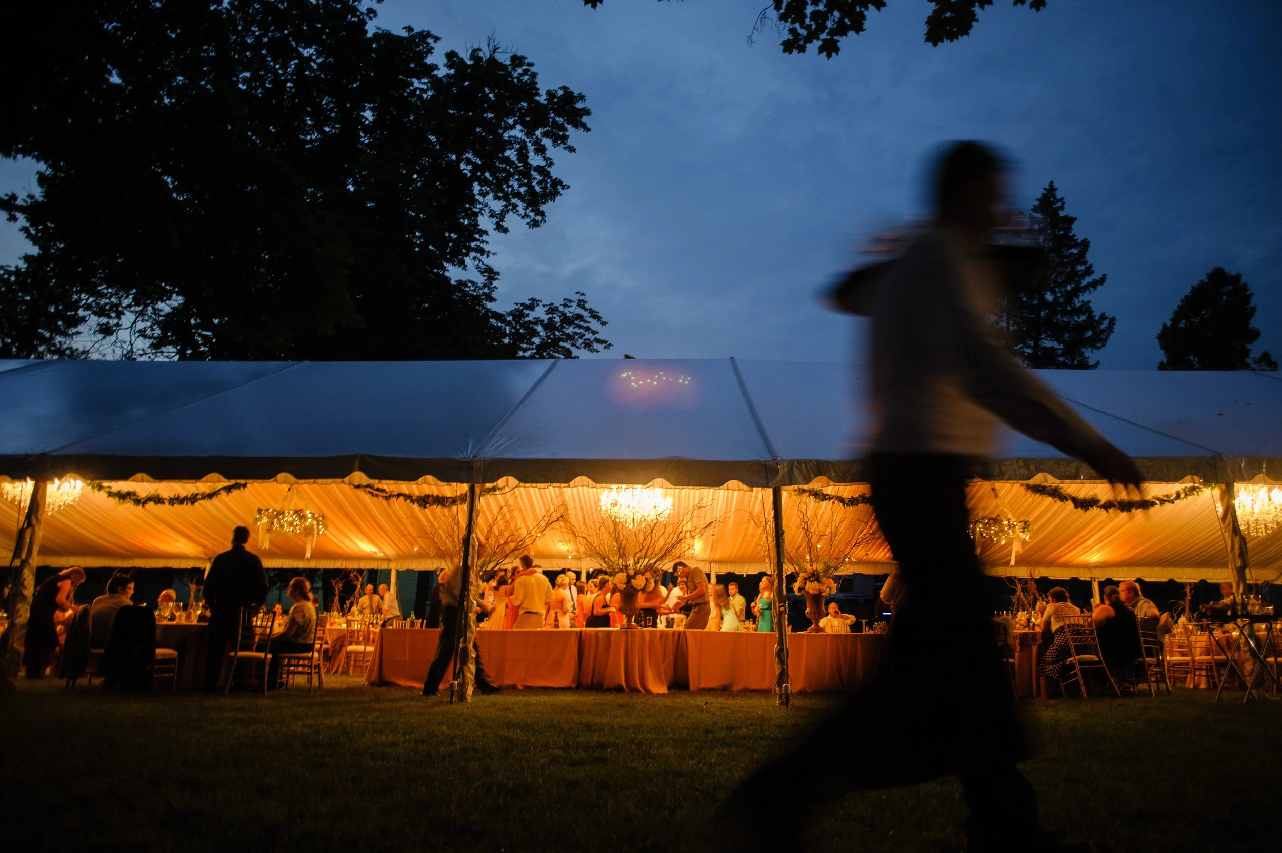 Blurred silhouette of a person walking past a warmly lit outdoor wedding reception under a large white tent at dusk, with trees and a cloudy sky in the background.