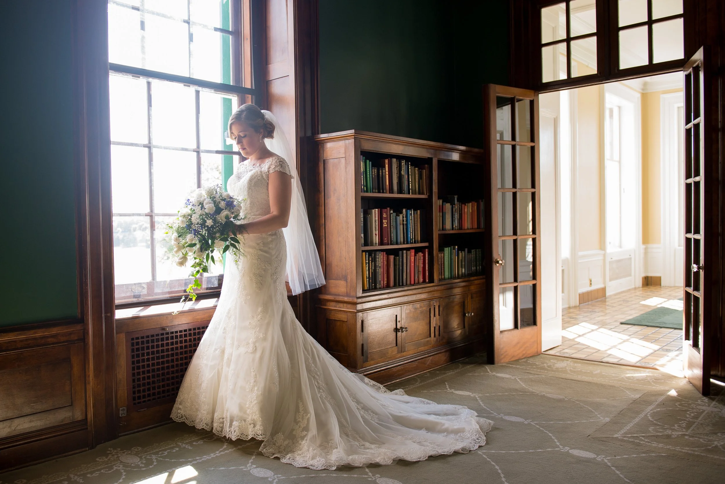 Bride in a white lace wedding dress holding a bouquet, standing by a large window in a room with dark green walls and wooden bookshelves, sunlight streaming in.